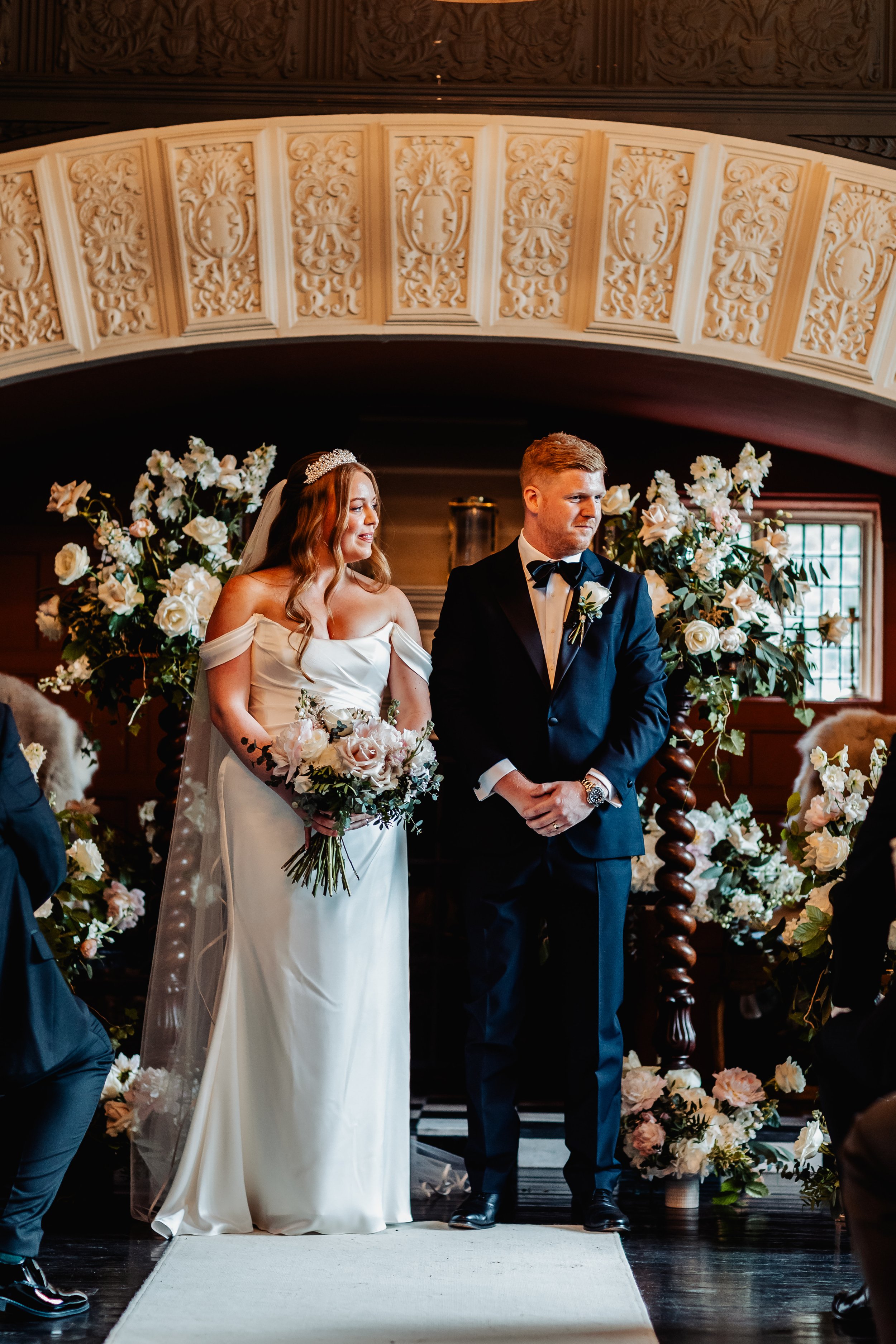 A bride and groom standing at the altar during their wedding ceremony, surrounded by floral arrangements, inside a decorated venue. East Yorkshire based wedding florals, handcrafted wedding flowers.