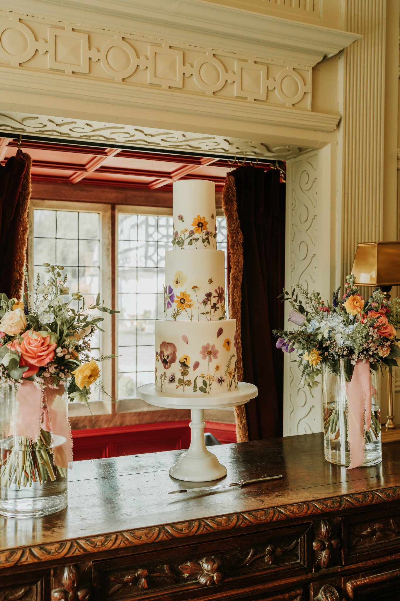 Four-tiered white wedding cake decorated with colorful hand-painted floral designs, placed on a white cake stand, flanked by two flower arrangements in glass vases, on a wooden table with intricate carvings, in a decorated indoor setting.