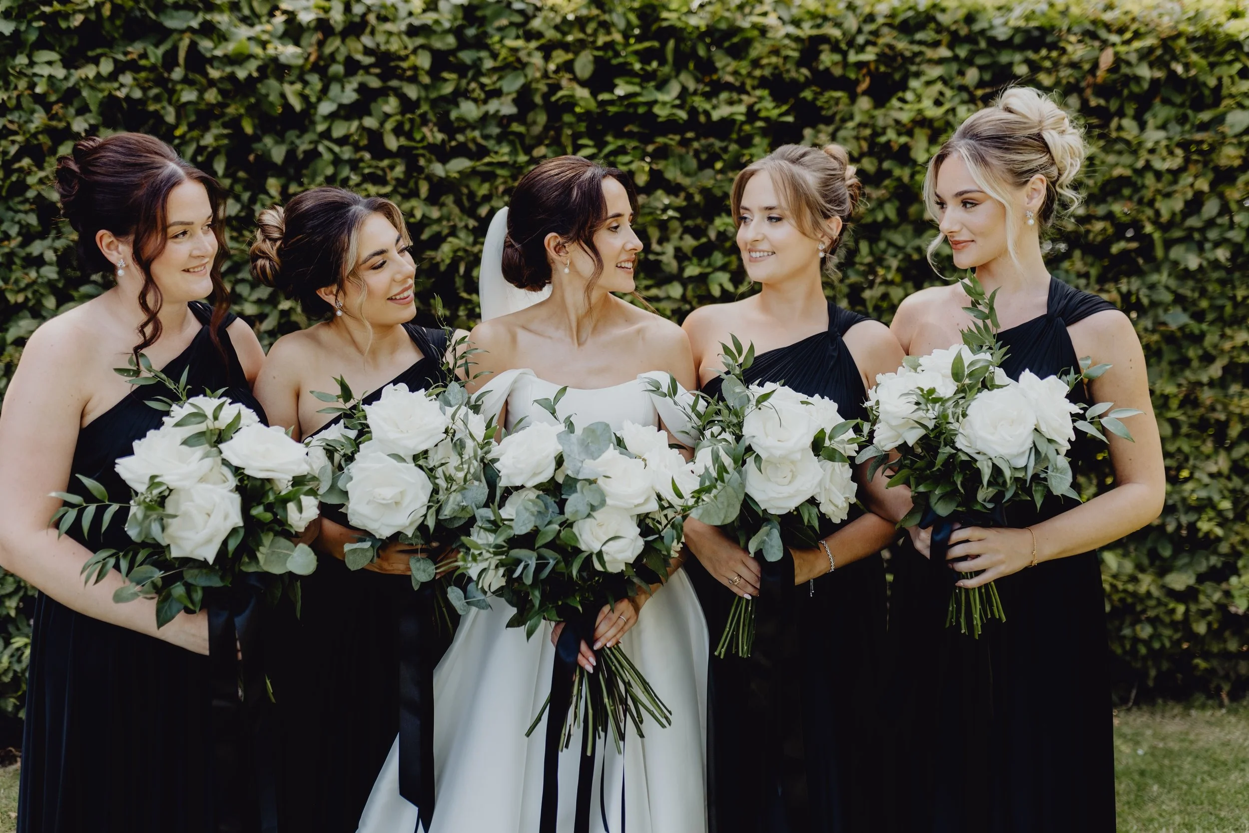 A bride in a white wedding dress standing among five bridesmaids in black dresses, all holding large bouquets of white roses and greenery, outdoors with a hedge background. East Yorkshire based wedding florals, handcrafted wedding flowers.