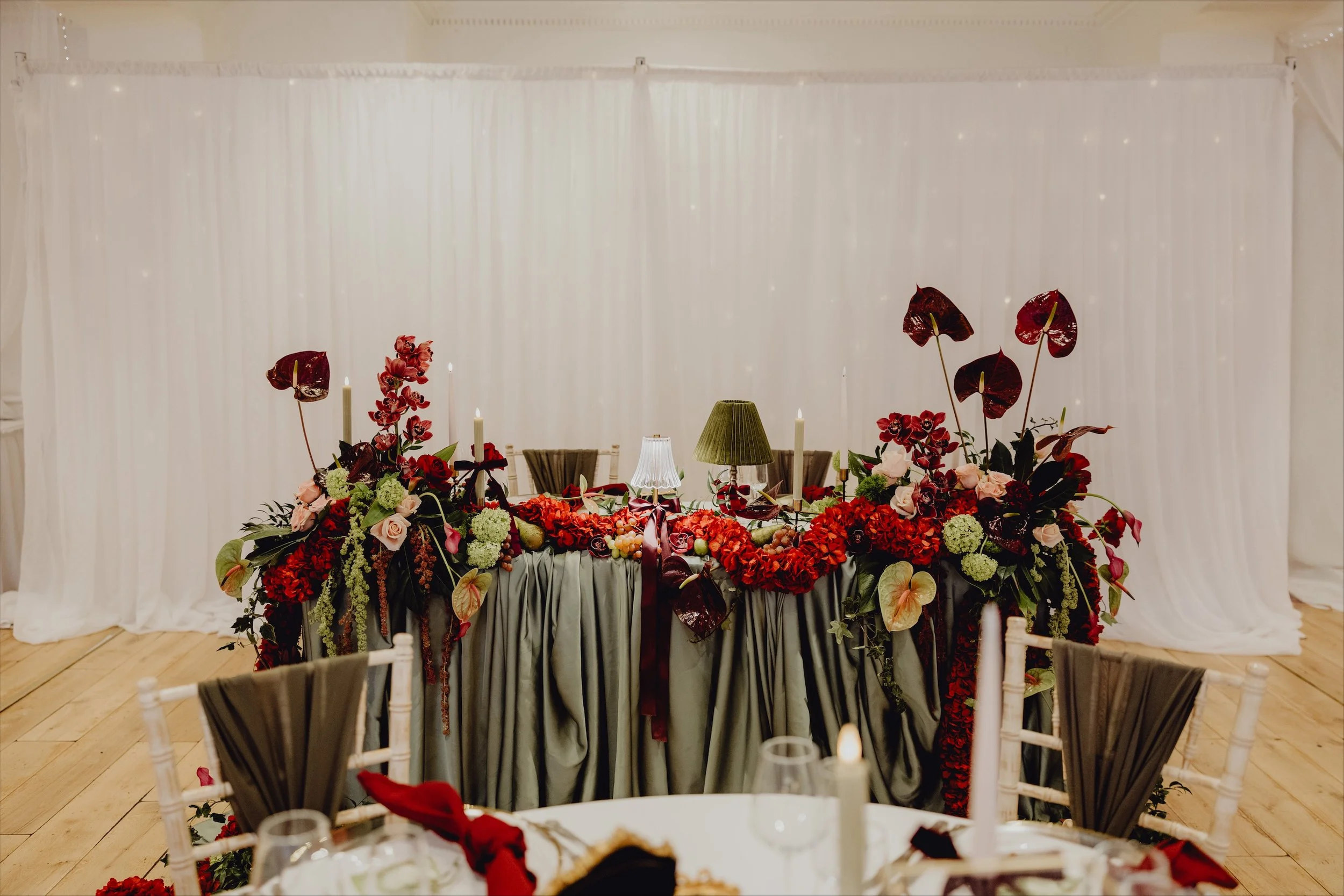 Wedding reception table decorated with red, pink, and green floral arrangements, white candles, and elegant tablecloths with a draped white backdrop.