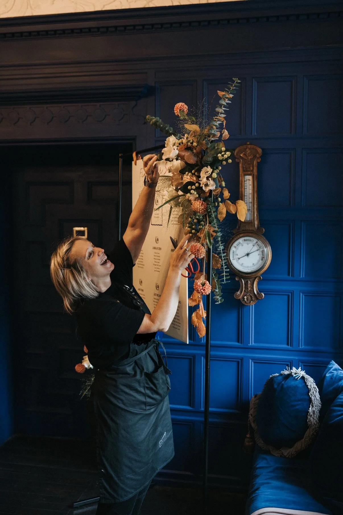 A woman arranging a large floral arrangement on a stand in a room with dark blue paneled walls, a vintage clock, and a blue sofa. East Yorkshire based wedding florals, handcrafted wedding flowers.