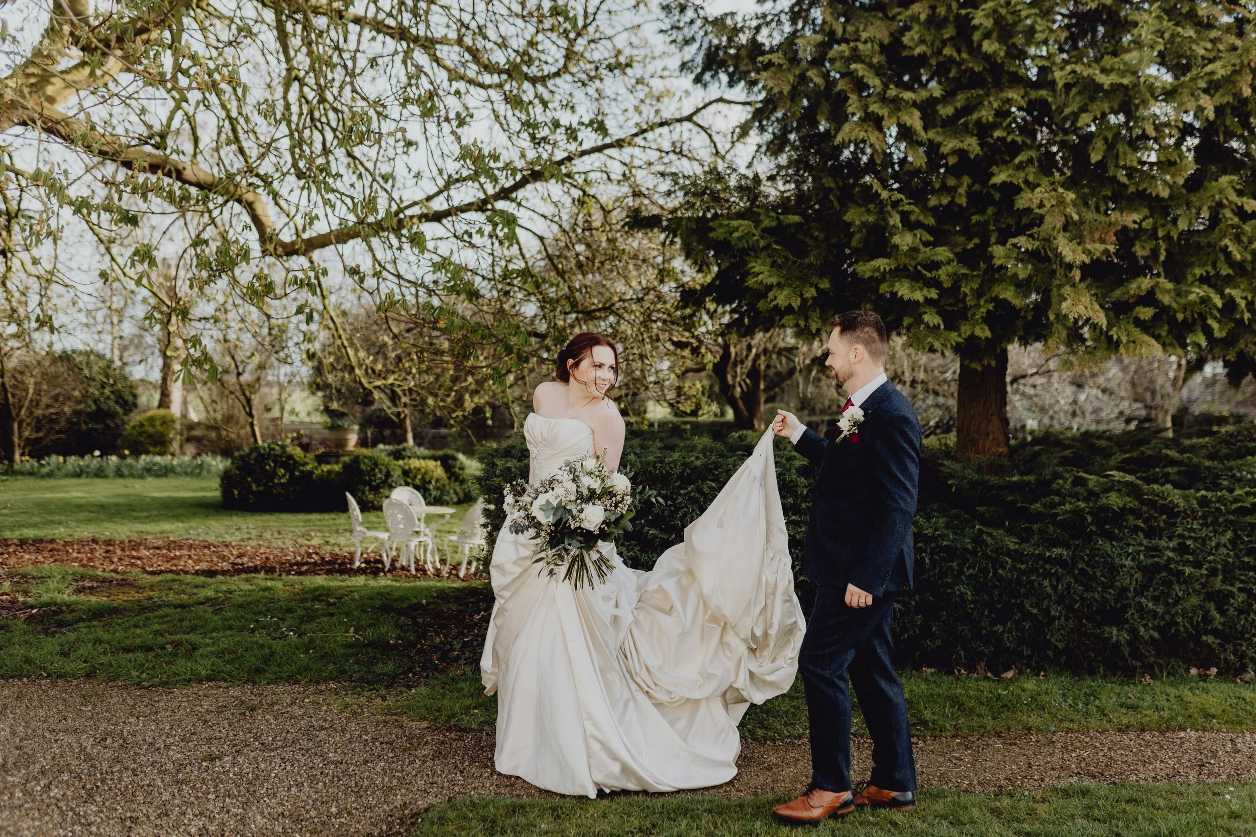 A bride and groom standing on a lawn in a garden, with the bride holding a bouquet and the groom holding the train of her wedding dress. They are smiling at each other under trees. East Yorkshire based wedding florals, handcrafted wedding flowers.