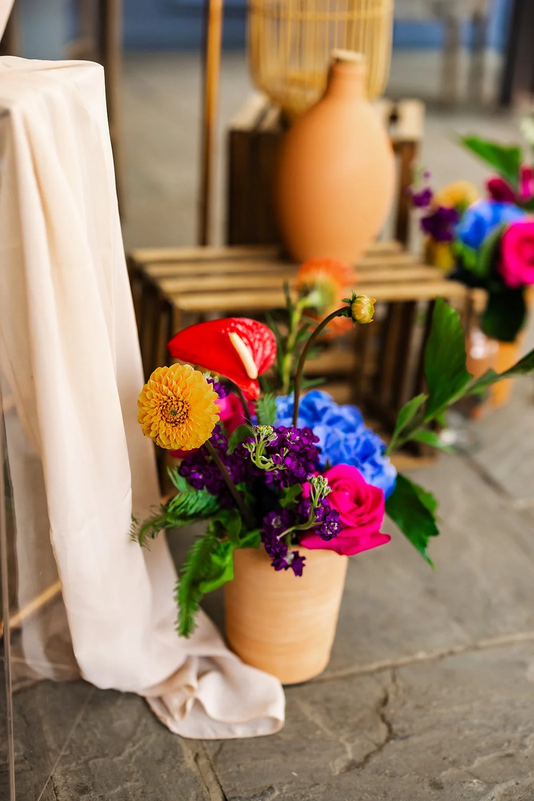 Colorful flower arrangement with pink roses, purple flowers, yellow flower, red anthurium, and green leaves in a beige clay pot, positioned on a wooden surface. East Yorkshire based wedding florals, handcrafted wedding flowers.