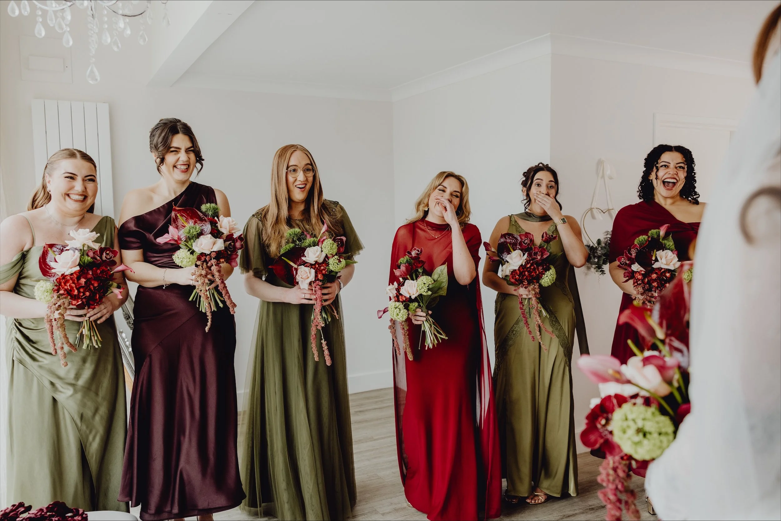 Group of six women in elegant dresses holding bouquets, smiling and laughing in a bright room.