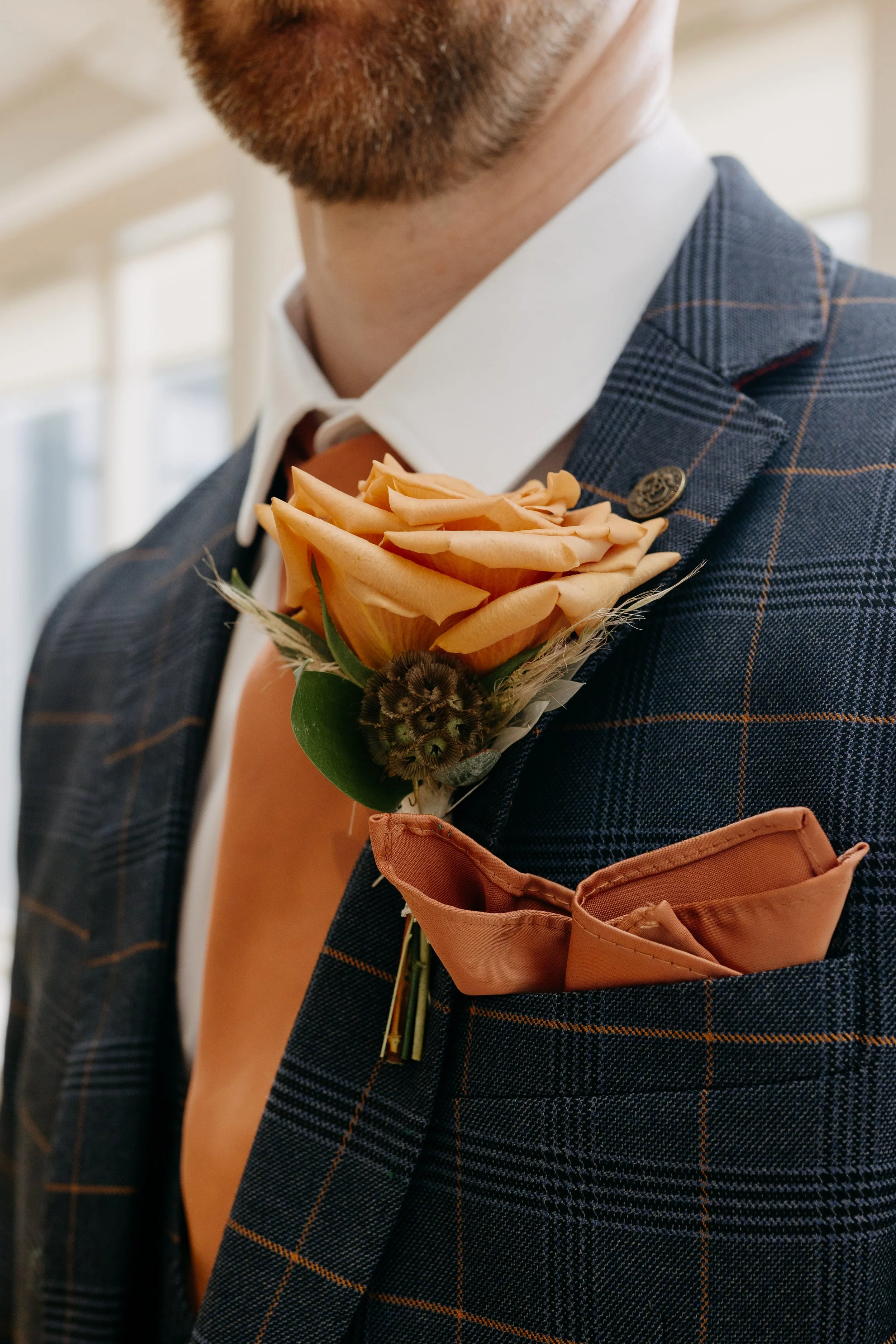 A man wearing a suit with a boutonniere made of a peach-colored rose, a green leaf, and a small brown fruit, with a pocket square in his pocket. East Yorkshire based wedding florals, handcrafted wedding flowers.