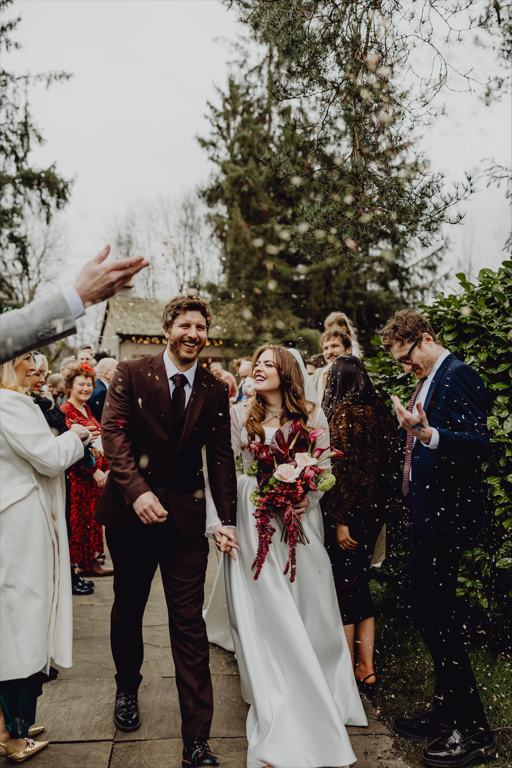A newlywed couple holding hands and smiling as they walk down a path during their wedding celebration, surrounded by friends and family outdoors, with trees and cloudy sky in the background.