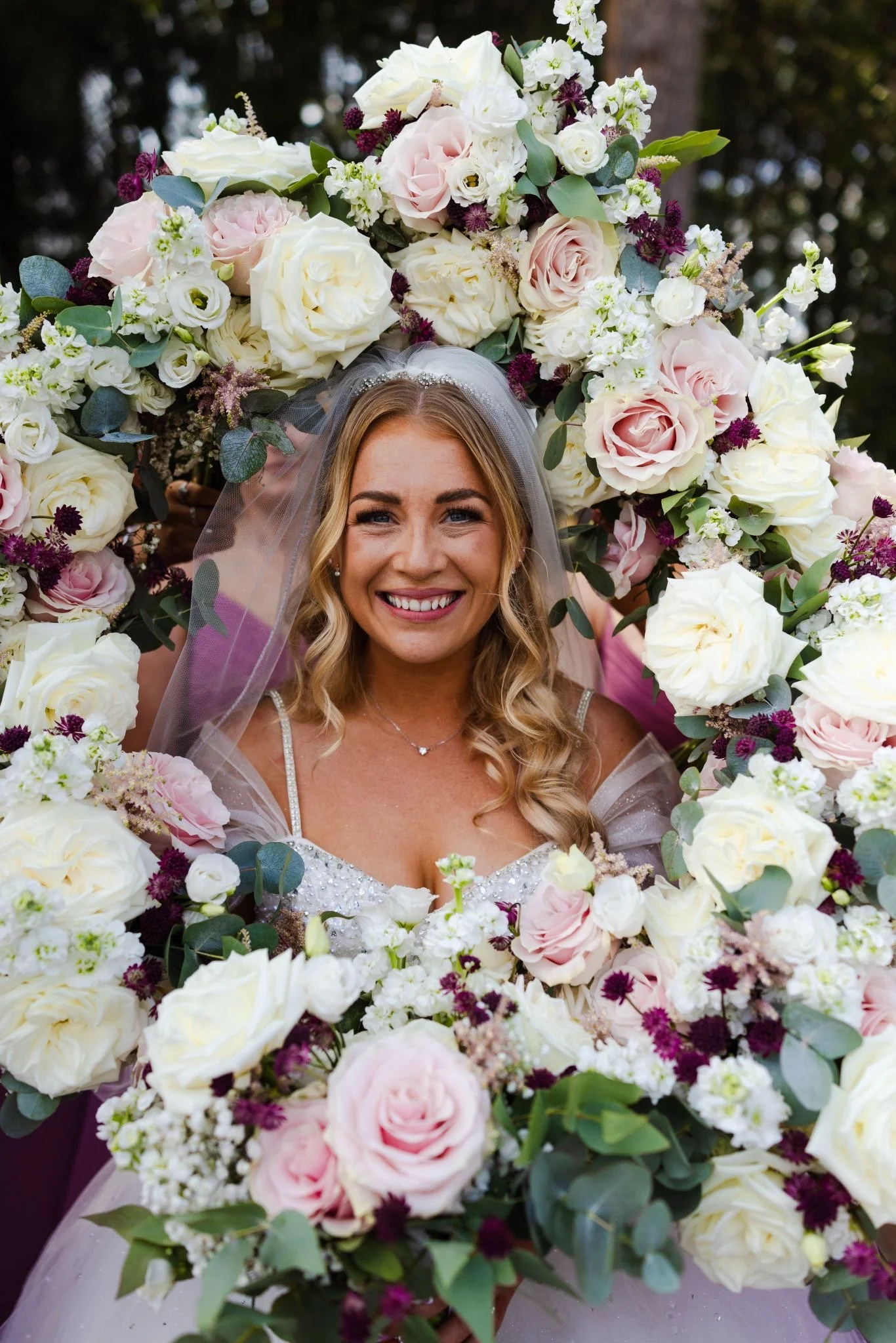 A smiling bride with blonde hair and a wedding dress, framed by a large floral arch of white, pink, and purple flowers and greenery. East Yorkshire based wedding florals, handcrafted wedding flowers.