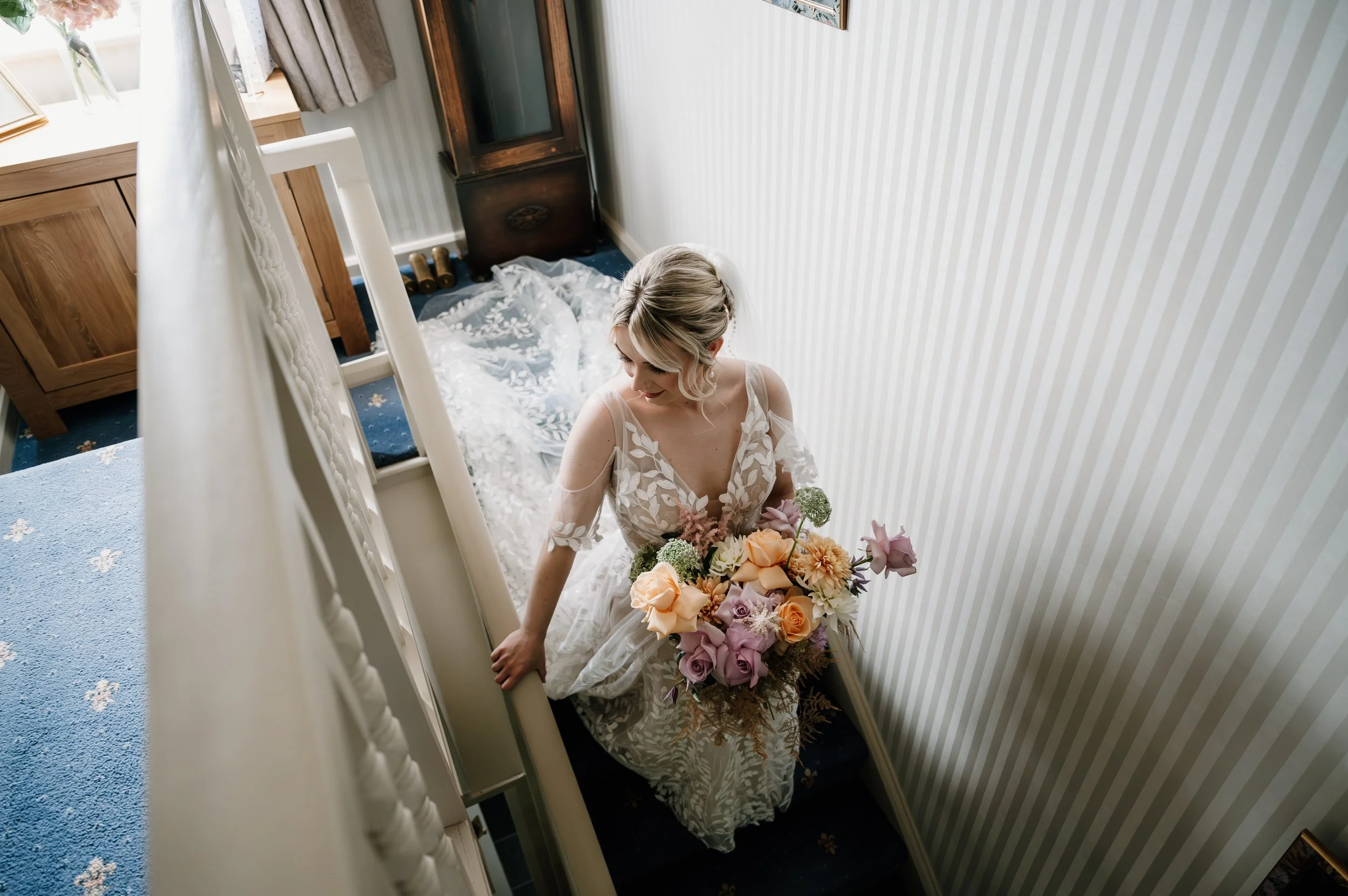 Bride standing on a staircase holding a bouquet of pastel roses and flowers, wearing a lace wedding dress with cold shoulder sleeves, in a cozy room with striped wallpaper and a bed with rumpled white bedding in the background.