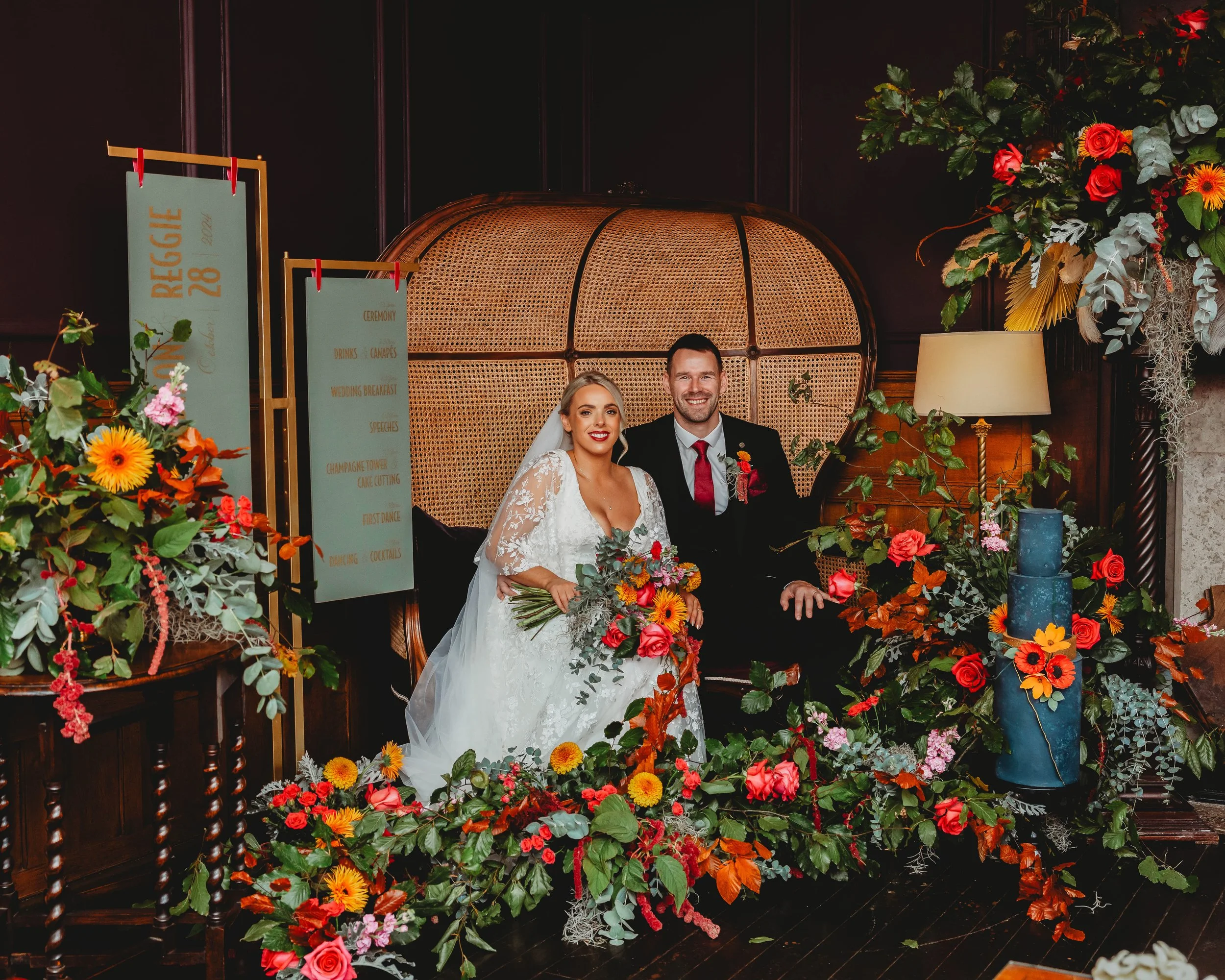A bride and groom sitting on a vintage-style sofa at their wedding reception with floral arrangements and a welcome sign in the background. East Yorkshire based wedding florals, handcrafted wedding flowers.