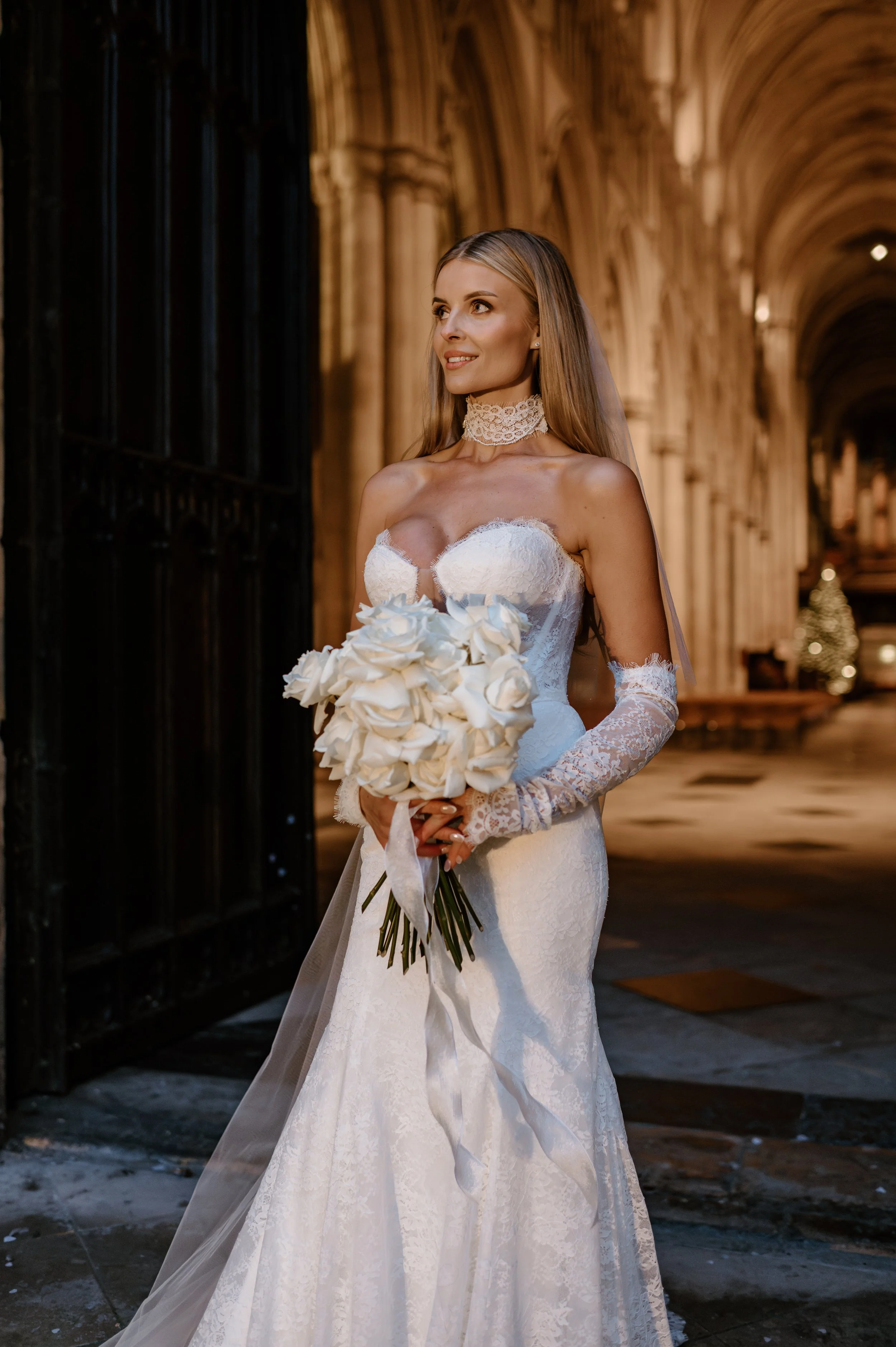 A bride in a white lace wedding gown holding a bouquet of white roses, standing in an arched stone hallway decorated with Christmas trees in the background. East Yorkshire based wedding florals, handcrafted wedding flowers.