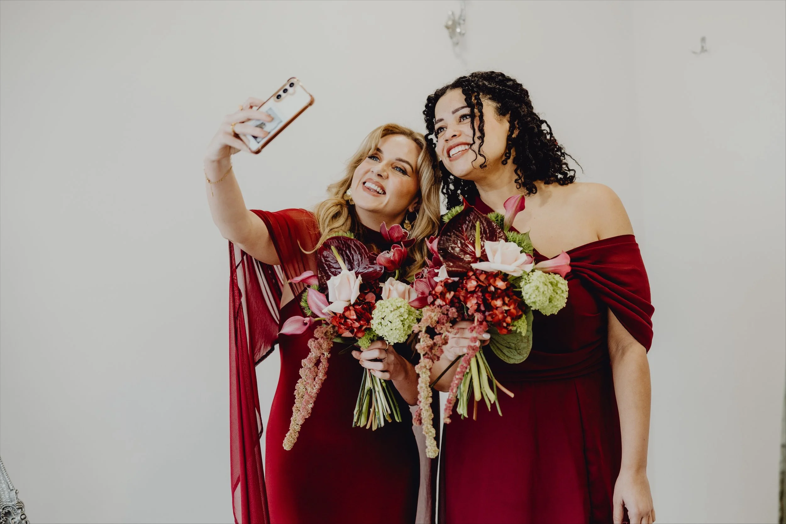 Two women in red dresses holding bouquets of flowers, taking a selfie together.