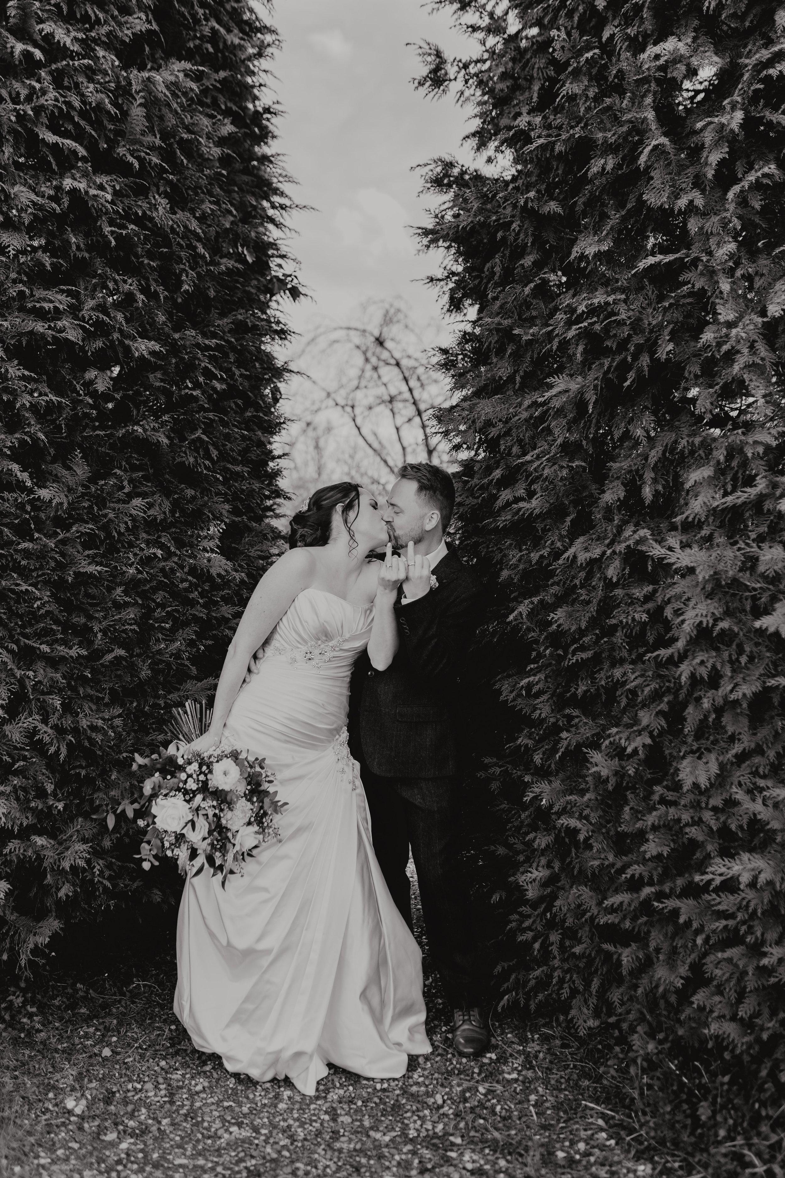 A black and white photo of a bride and groom sharing a kiss between tall evergreen trees, with the bride holding a large bouquet. East Yorkshire based wedding florals, handcrafted wedding flowers.