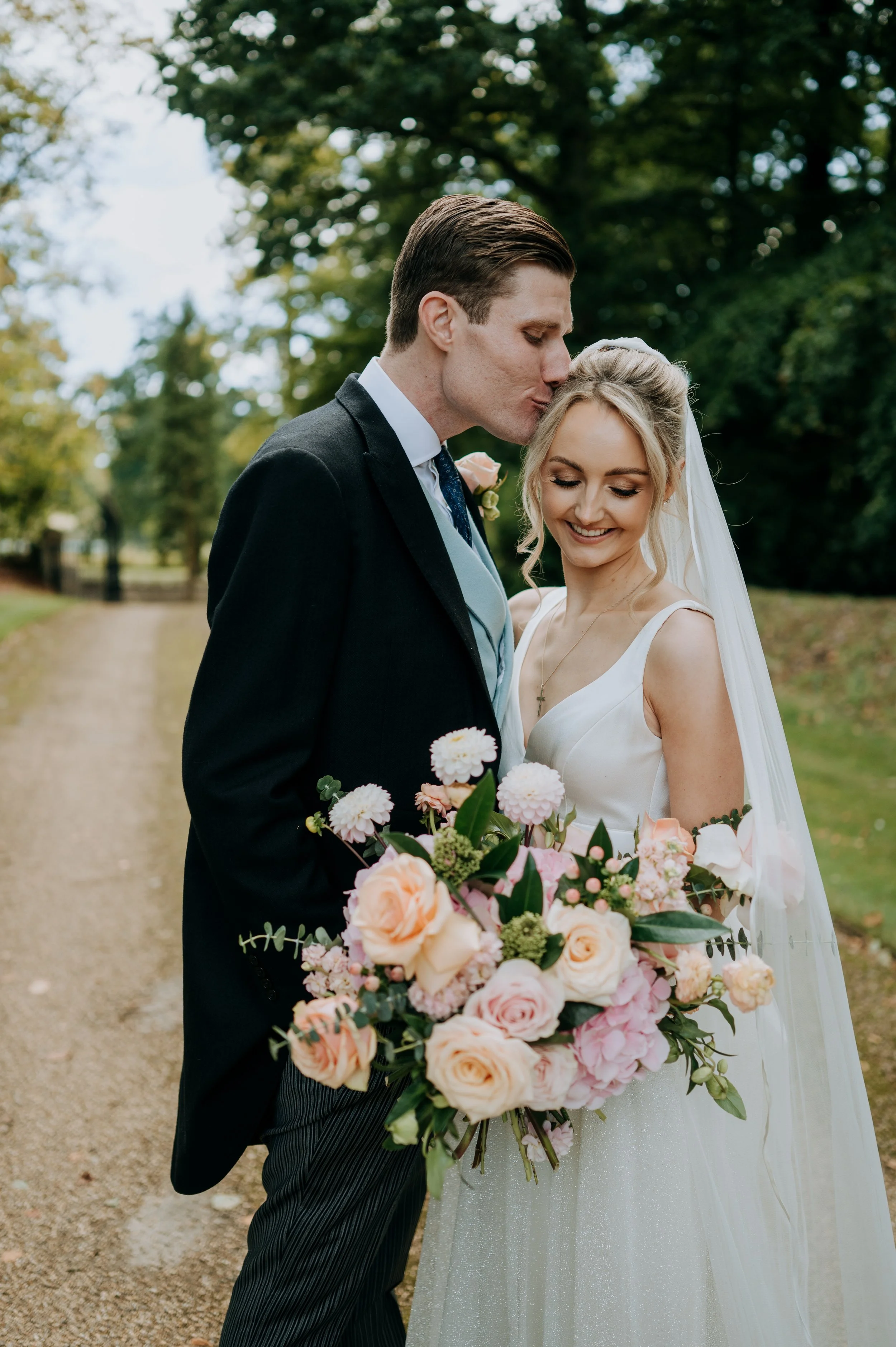 A newlywed couple stands outdoors on a tree-lined path, with the groom kissing the bride's forehead. The bride wears a white wedding dress and holds a large bouquet of pink and peach flowers, while the groom is dressed in a black suit.