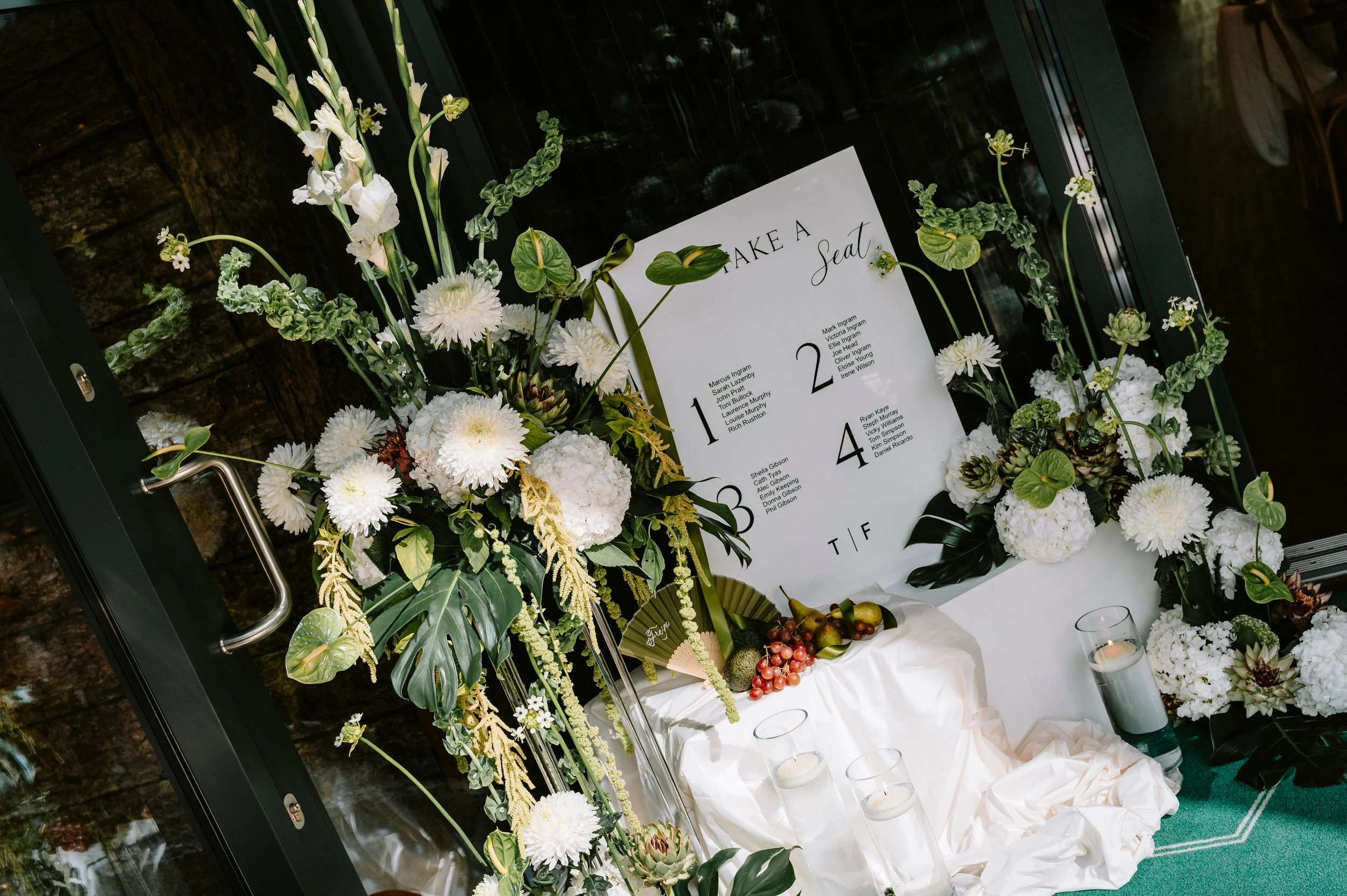 Wedding seating chart displayed among white flowers, candles in glass holders, and greenery.