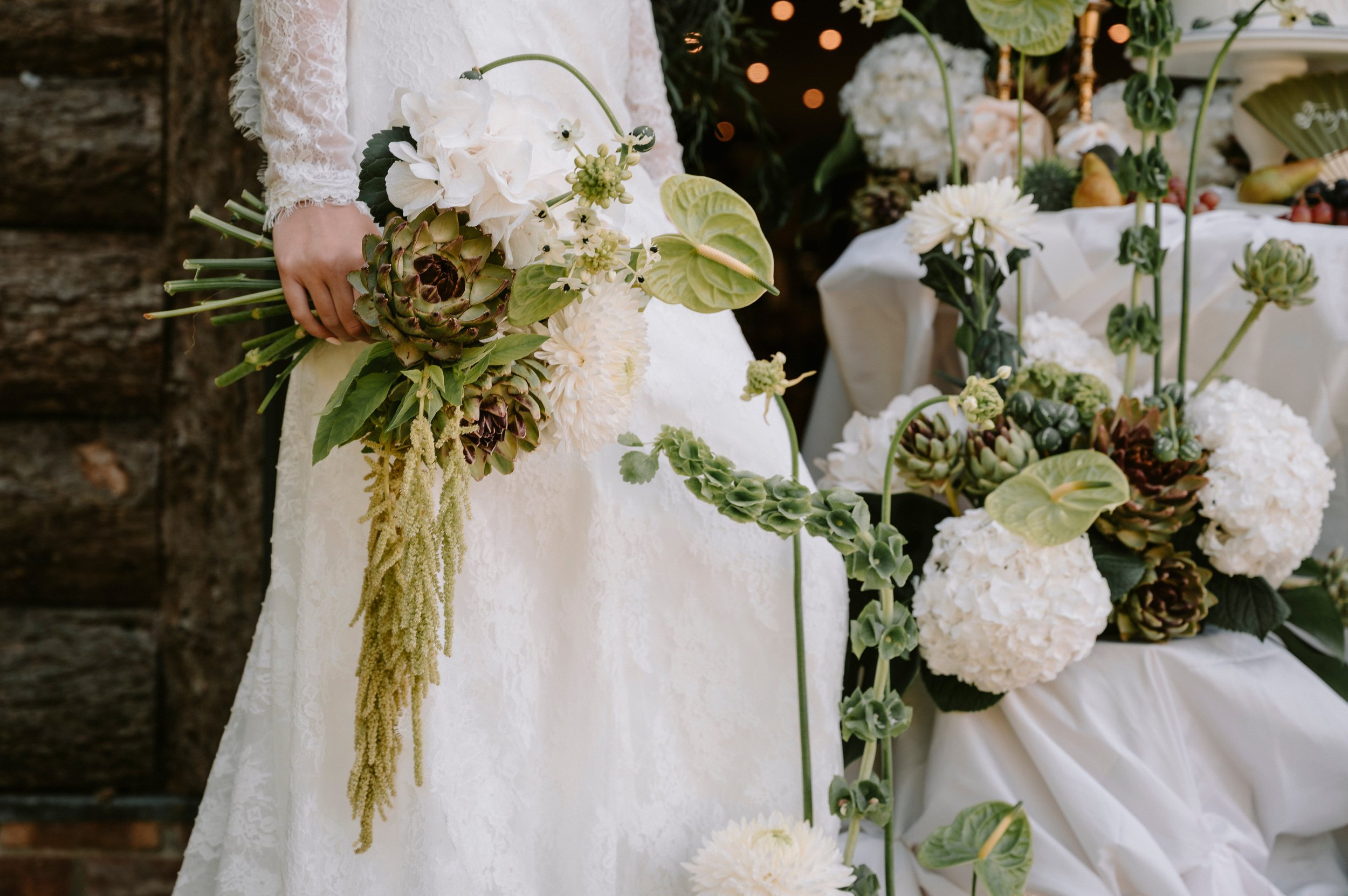 A person in a white lace dress holding a cascading bouquet of white and green flowers and succulents at a wedding or event with a decorated table in the background.