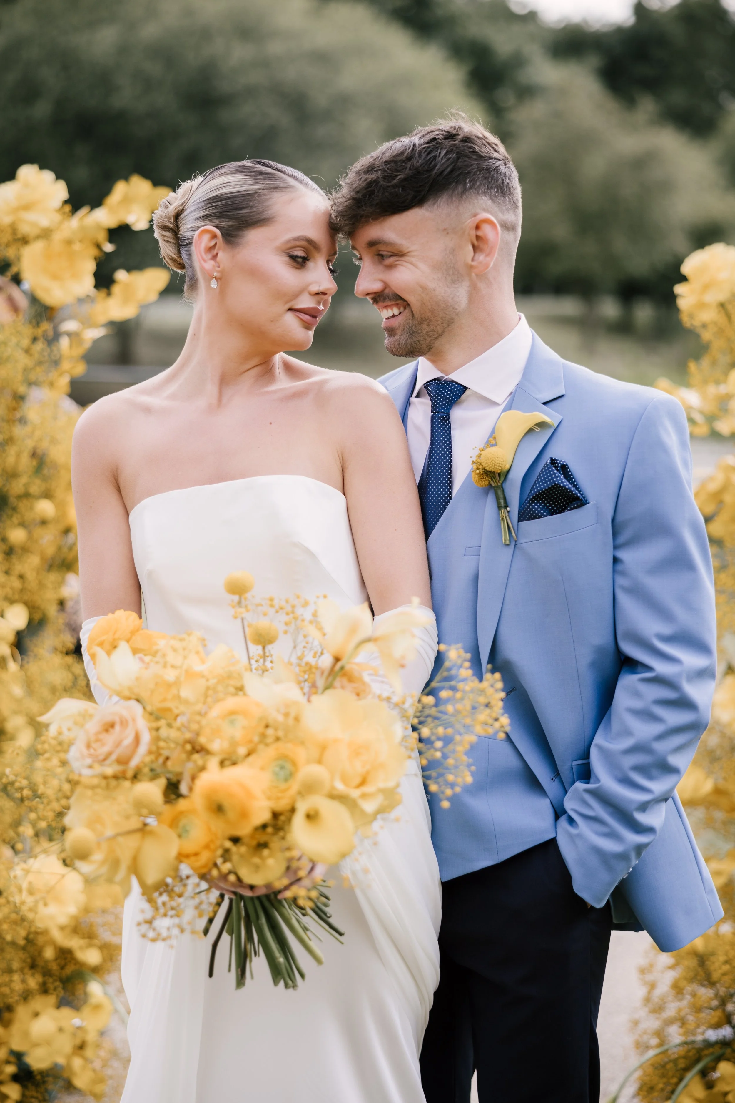A couple in wedding attire standing close together outdoors with yellow flowers around them, smiling and looking into each other's eyes. The bride holds a yellow floral bouquet, and the groom wears a light blue suit with a boutonnière.