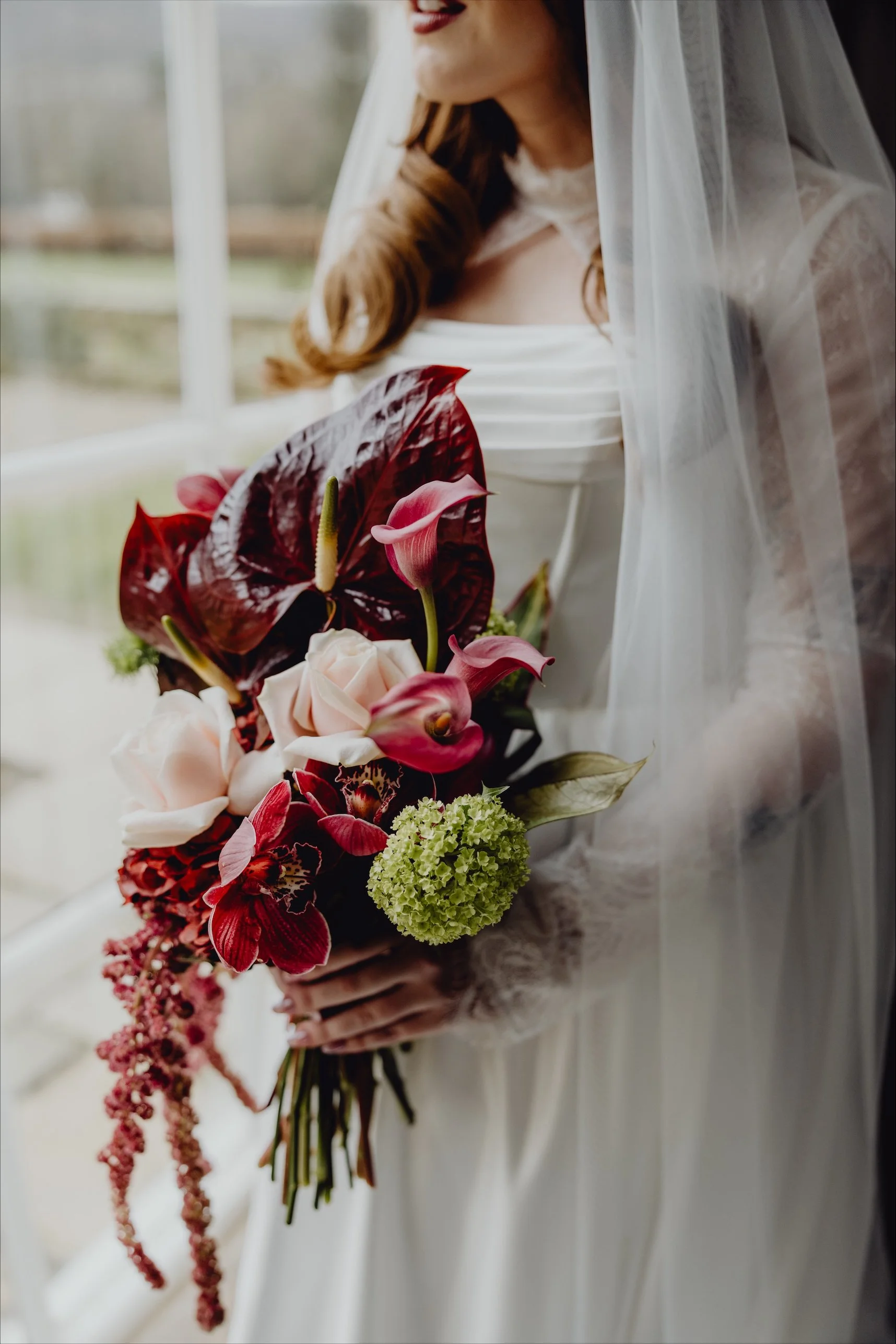 A bride holding a bouquet of pink and red flowers, including calla lilies, orchids, and hydrangeas, in front of a window.