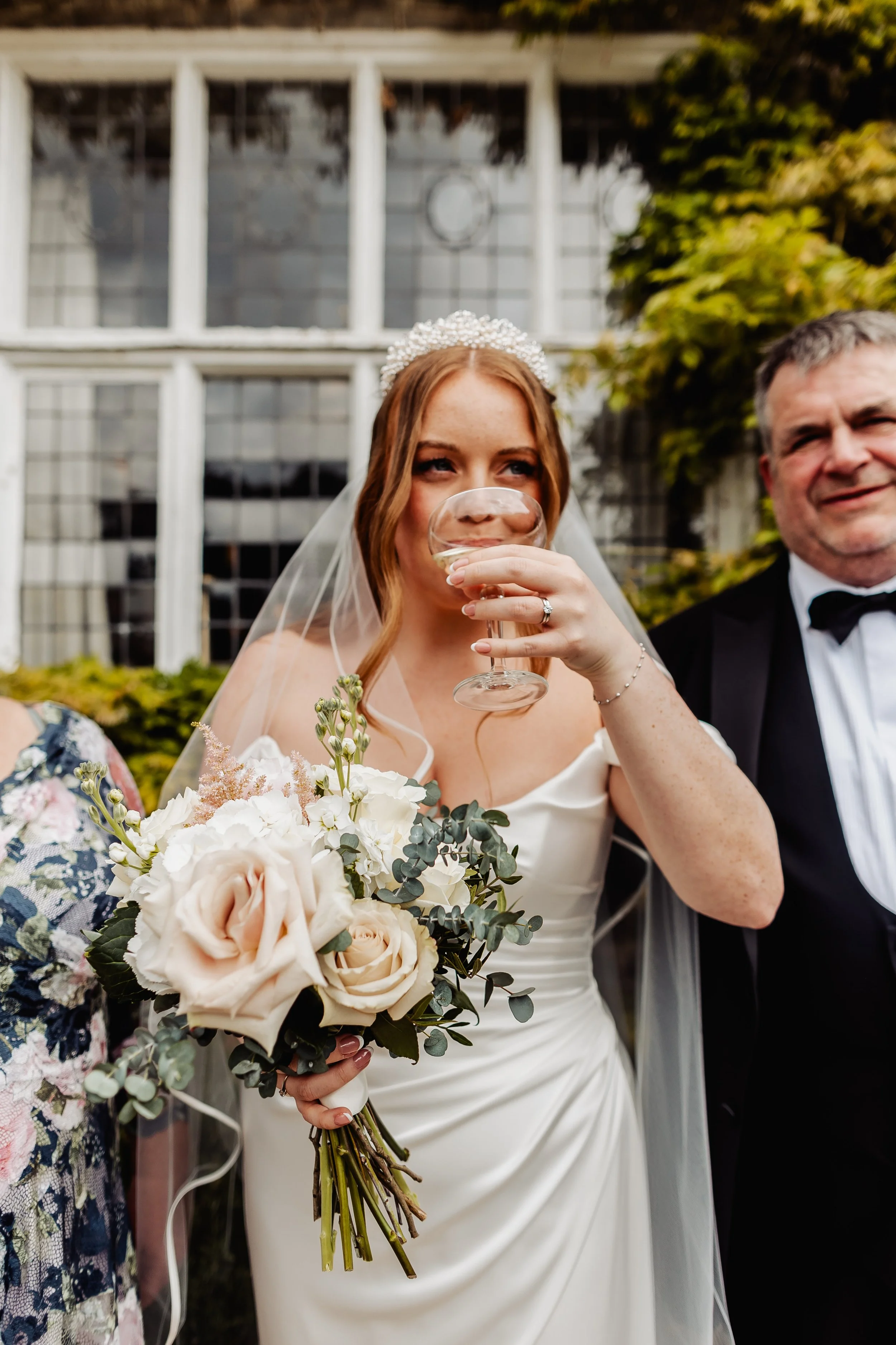 A bride with red hair in a white wedding gown and veil, holding a bouquet of cream and blush roses and eucalyptus, drinking from a champagne glass, standing outdoors next to a man in a tuxedo during a wedding celebration.