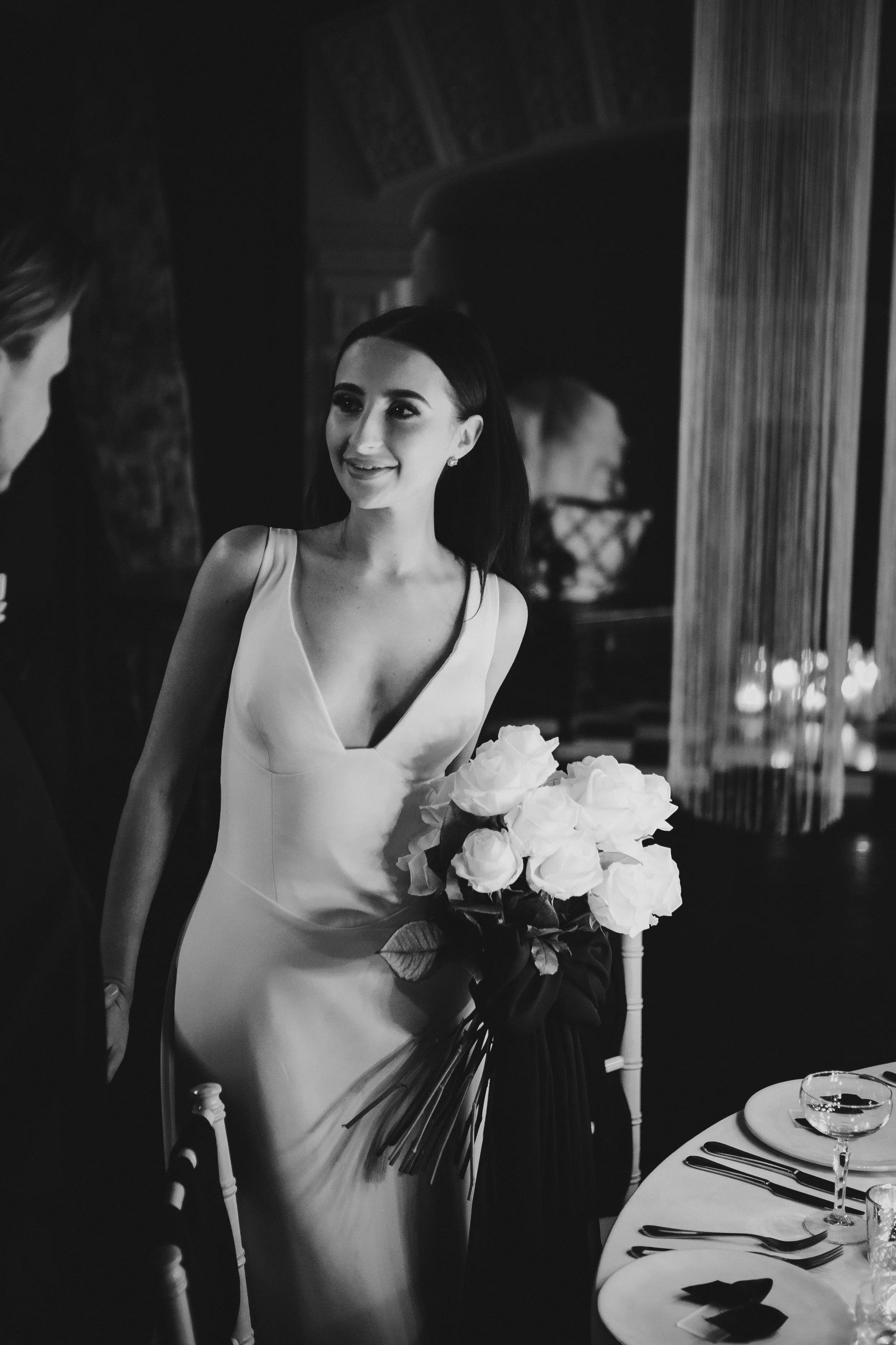 A woman in a wedding dress holding a bouquet of white roses, smiling and talking to a man at a wedding reception.East Yorkshire based wedding florals, handcrafted wedding flowers.