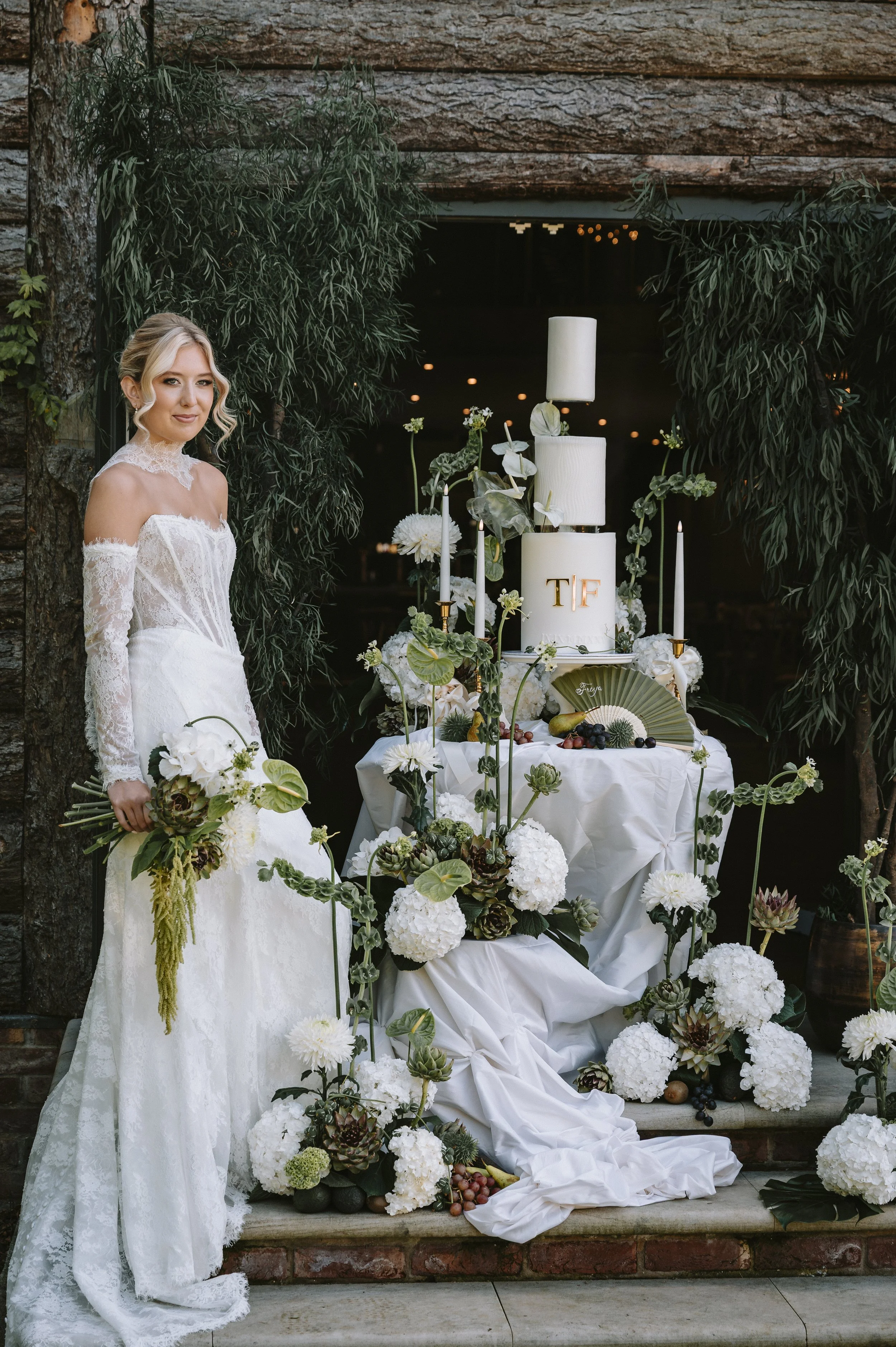A bride in a white lace wedding gown standing beside a decorated wedding cake with white flowers and greenery at an outdoor wedding venue. East Yorkshire based wedding florals, handcrafted wedding flowers.