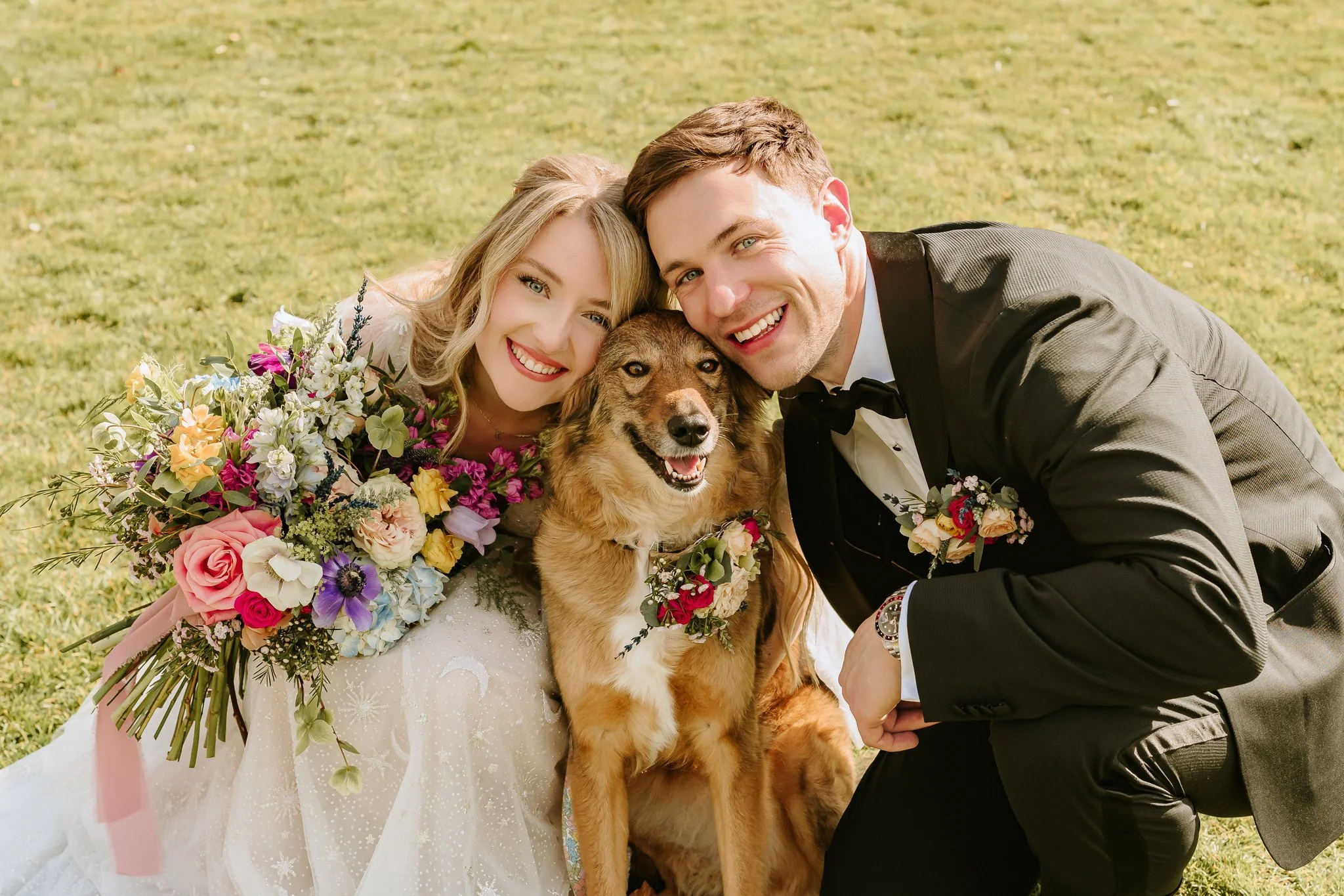 A happy bride and groom with a dog, posing outdoors on grass during their wedding. East Yorkshire based wedding florals, handcrafted wedding flowers.