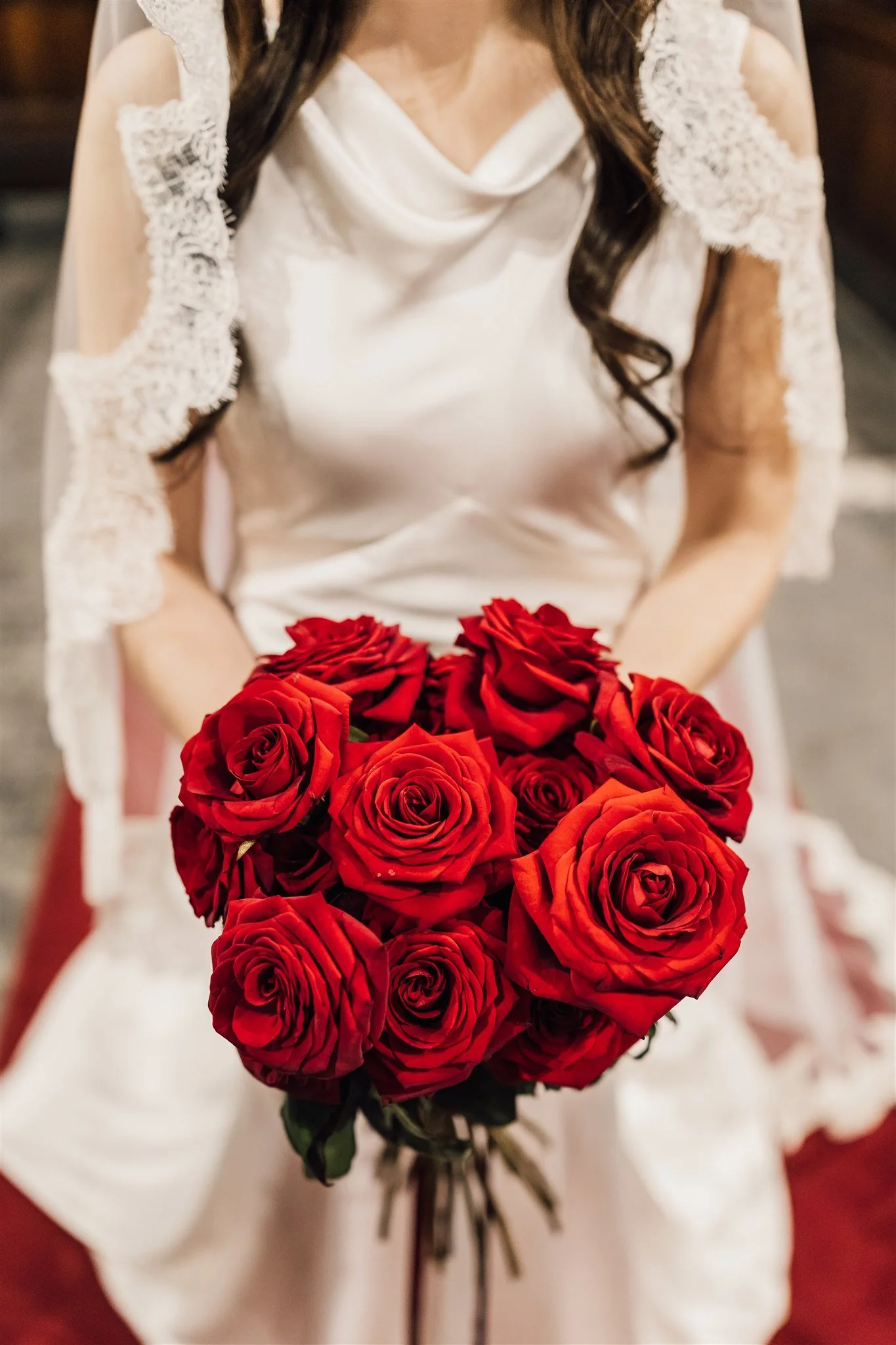 A woman in a white dress with lace details on the sleeves is holding a large bouquet of red roses. East Yorkshire based wedding florals, handcrafted wedding flowers.