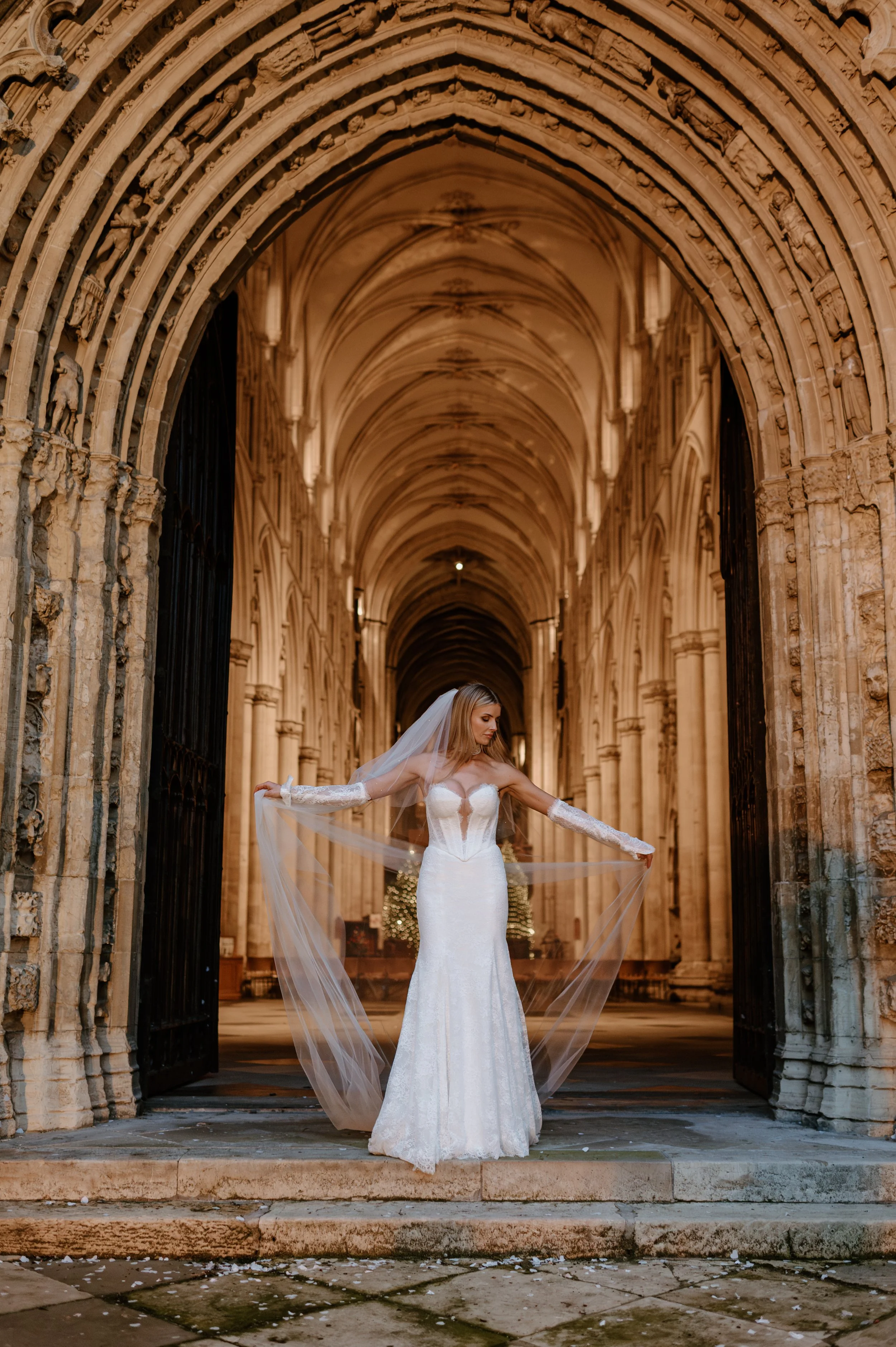 A bride in a white wedding gown with a veil standing in an ornate stone archway of a historic church or cathedral, holding the edges of her veil and posing with a serene expression. East Yorkshire based wedding florals, handcrafted wedding flowers.