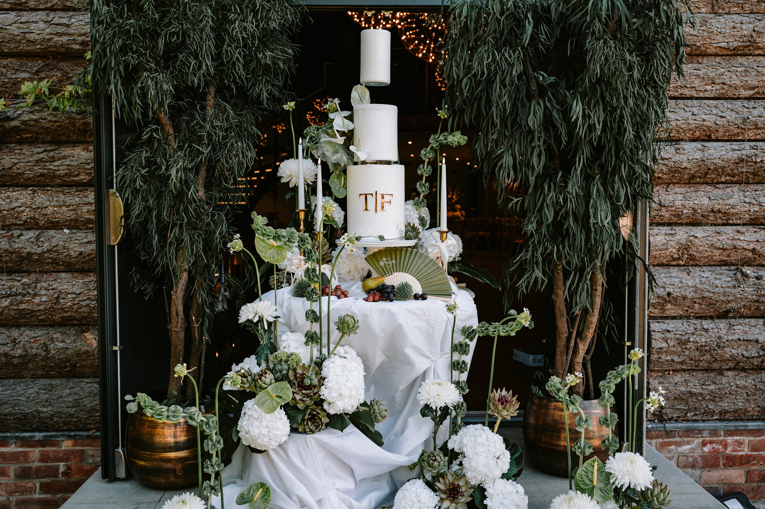 A wedding cake with white fondant, gold letters 'TIF,' surrounded by white flowers, candles, and greenery, set on a table draped with white fabric. The setup is framed by potted plants and a rustic brick and wood wall.