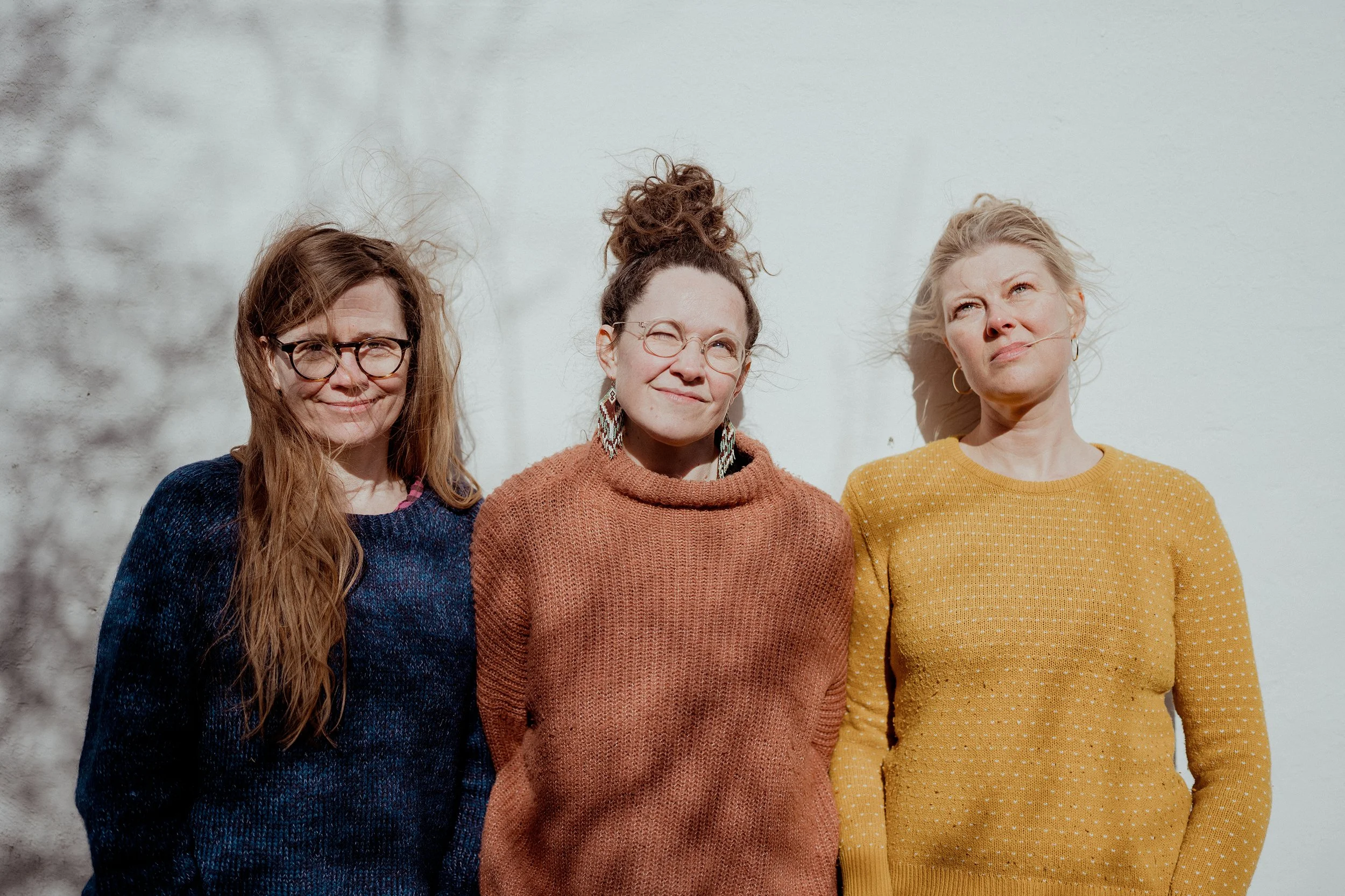 Tre kvinder står ude foran en hvid væg og nyder solen, hvoraf den midterste har kurvet hår oppe i en knold, den venstre har brunt, langt hår og briller, og den højre har blond, fritflyvende hår og gule tøj.