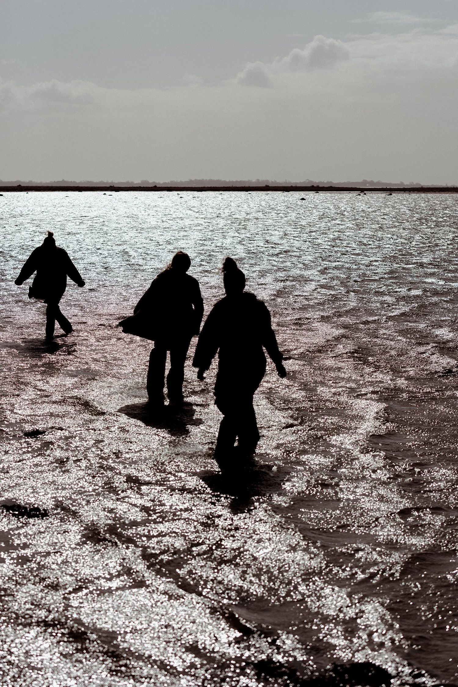 Silhuetter af fire mennesker, der går i vandet ved stranden, med sollys, der reflekterer på vandet, og en åben himmel med spredte skyer i baggrunden.