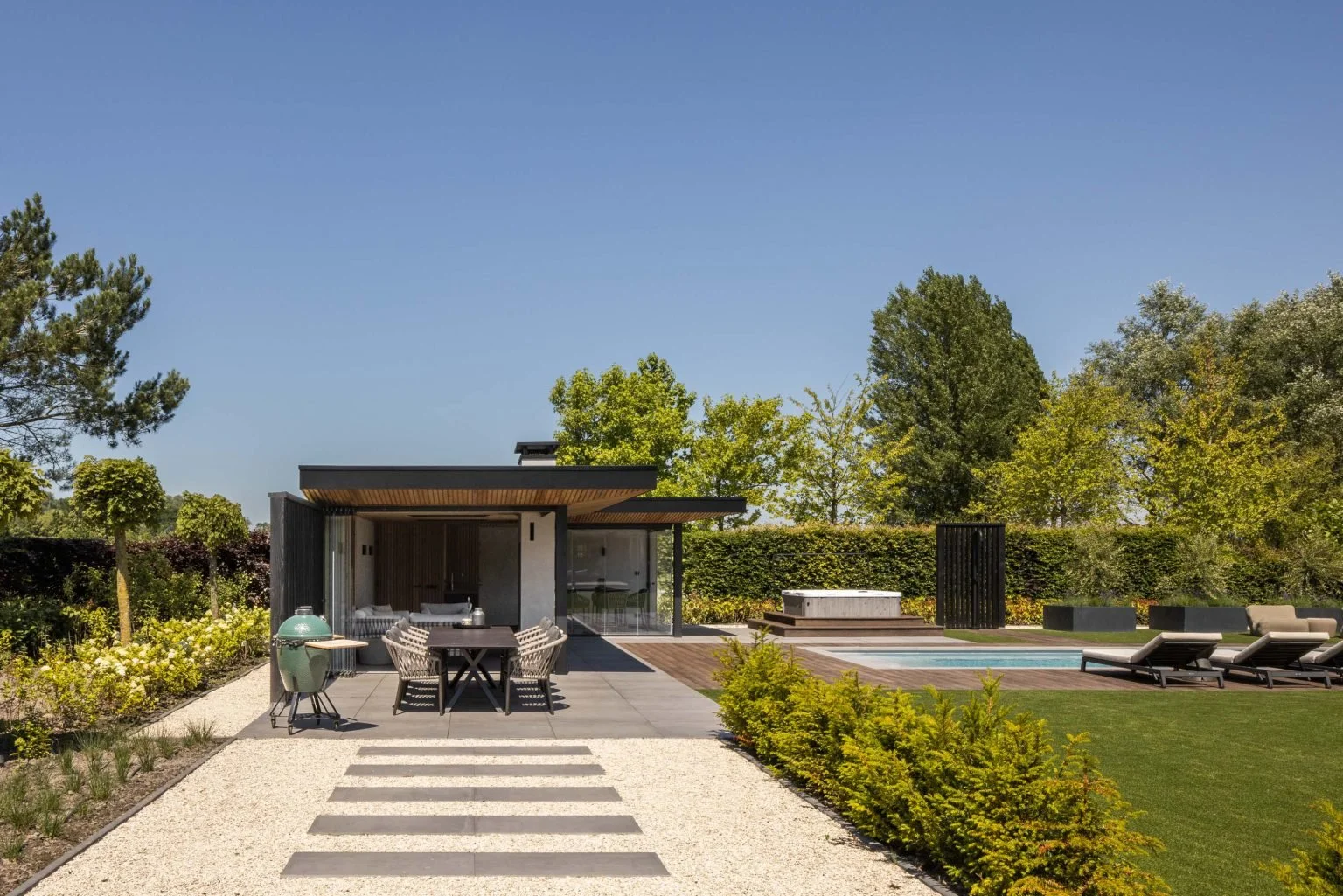 Een moderne tuin met een terras, pergola, zwembad en ligstoelen, omgeven door groen bomen en struiken onder een heldere blauwe hemel.