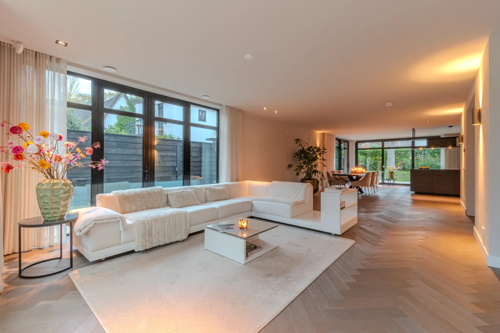 Luxe zitkamer met grote witte hoekbank, vloerkleed, bijzettafel met vaas met kleurrijke bloemen, grote ramen met gordijnen, eettafel met stoelen en keuken in de achtergrond, houten vloeren, zachte verlichting.