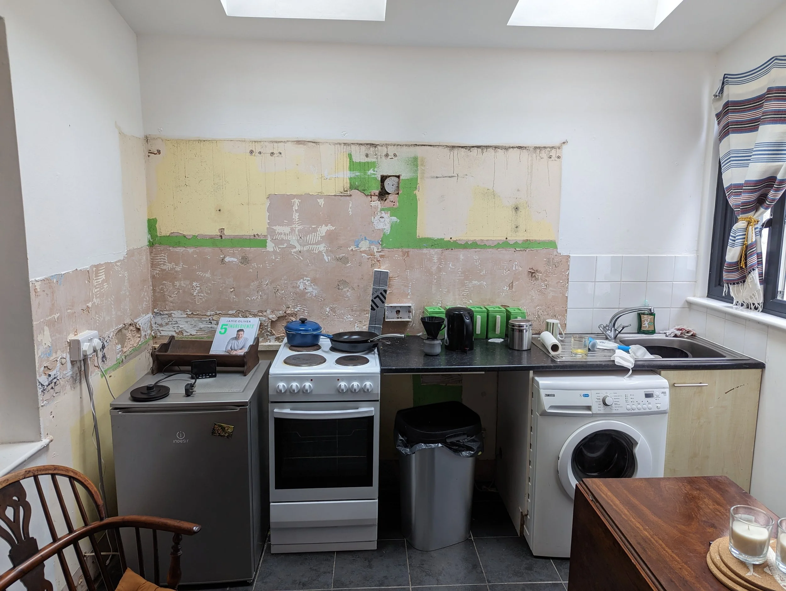 A kitchen under renovation with a partially removed wall, showing exposed patches of plaster and electrical outlets.