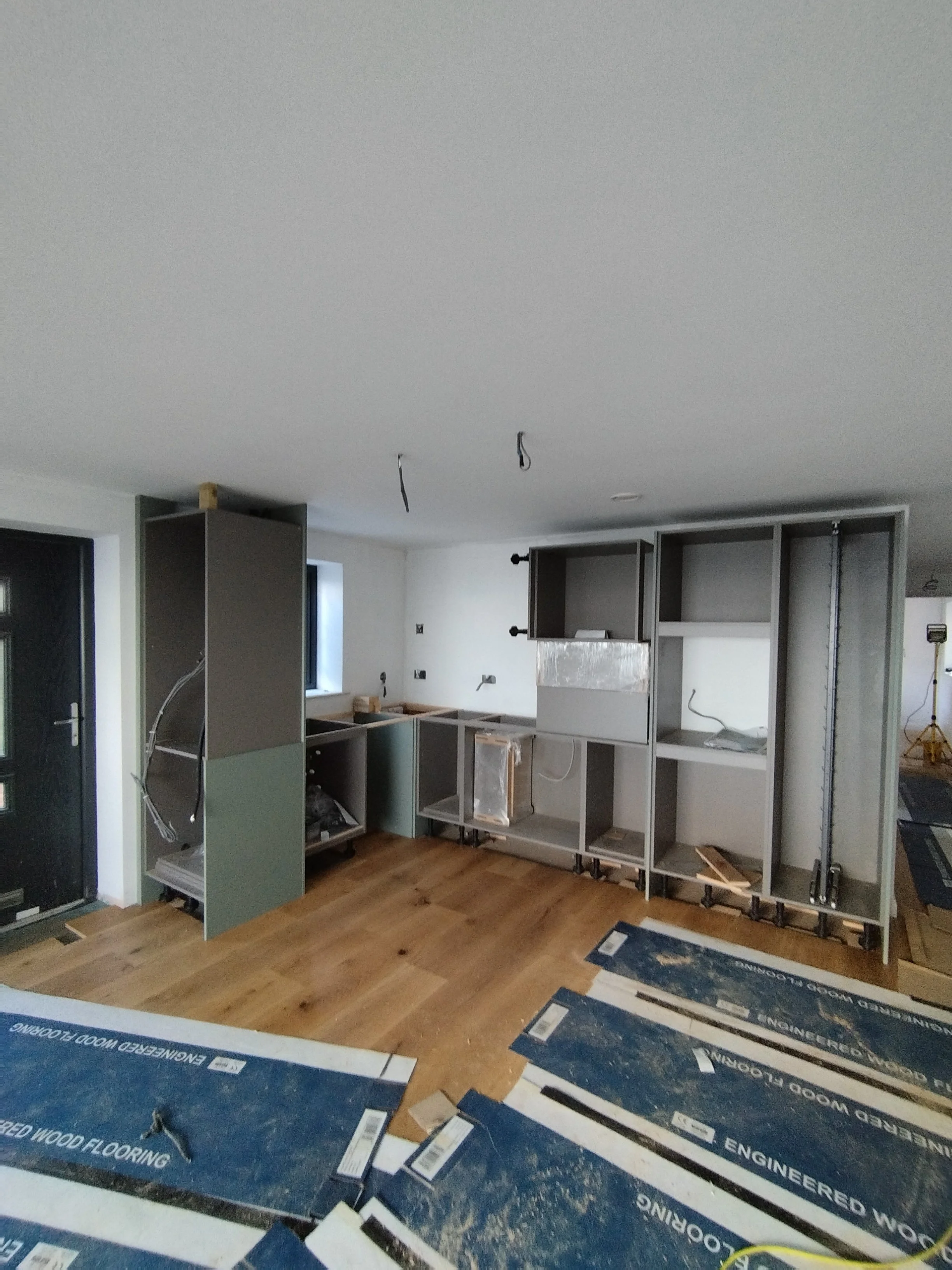 Kitchen under construction with unfinished cabinets, wiring hanging from the ceiling, and wood flooring partially installed.