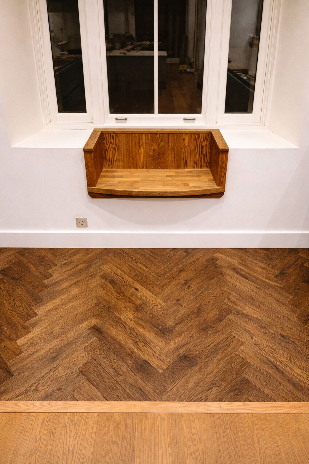 A wooden window seat below a large window in a room with hardwood herringbone flooring.
