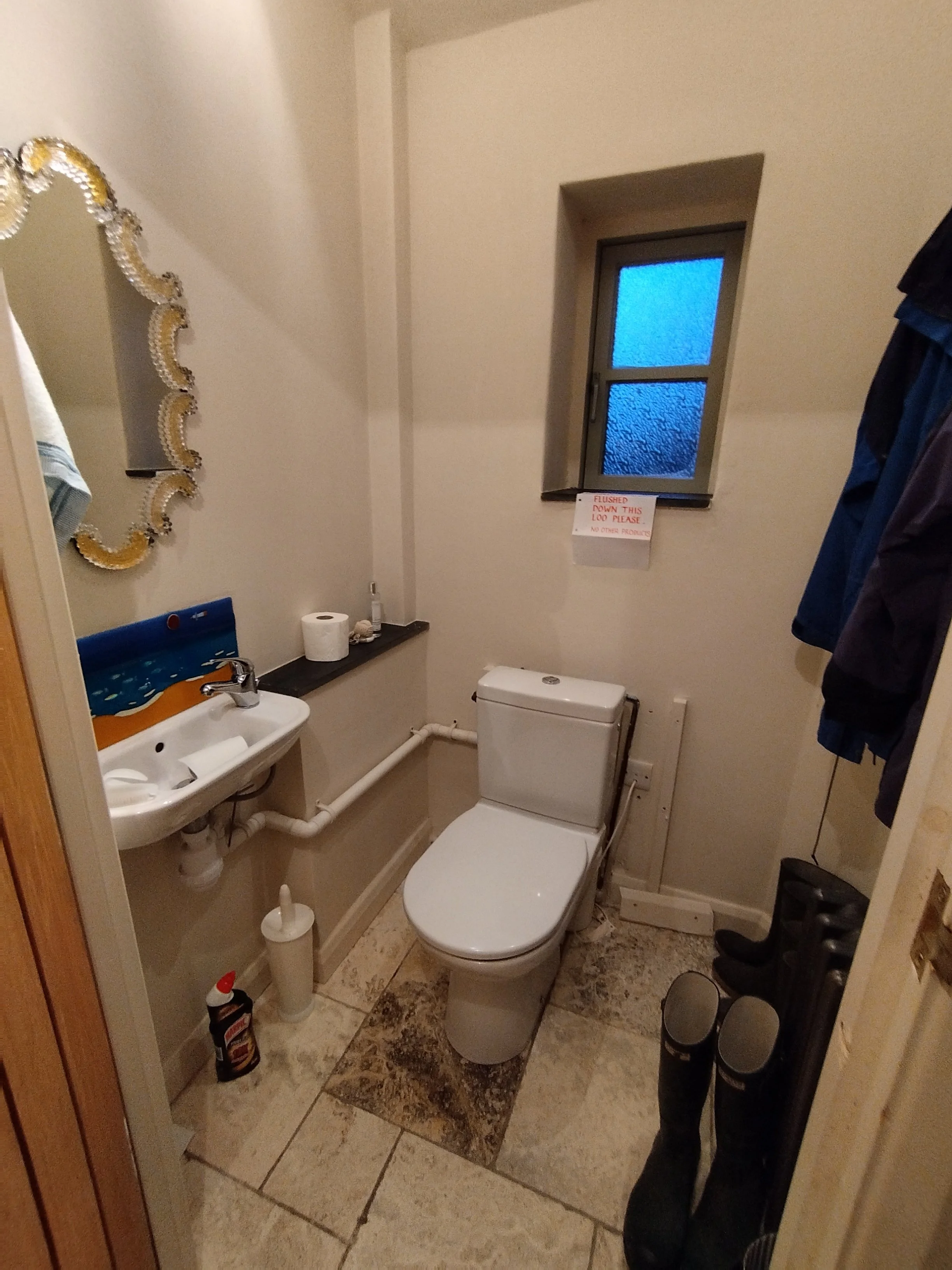 Small bathroom with a toilet, a small sink with a mirror, a window with a sign, and some shoes and cleaning supplies.