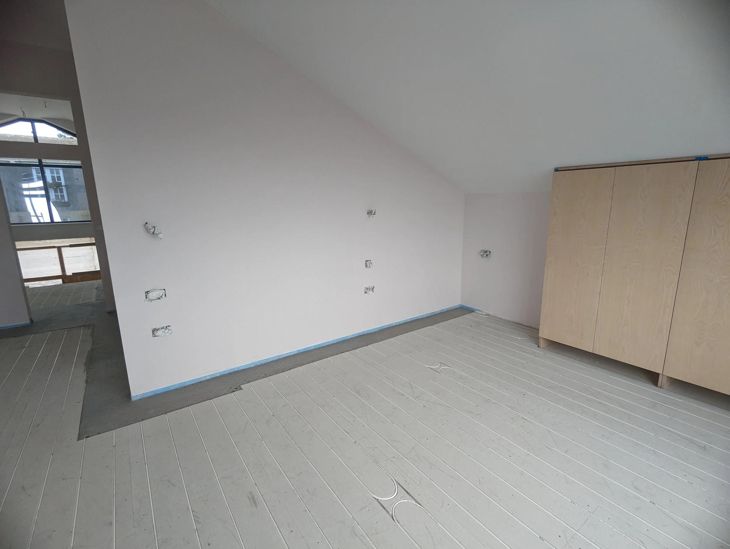 An unfinished room with white walls, some electrical wires and outlets, a wooden wardrobe on the right, and light-colored flooring with visible seams.