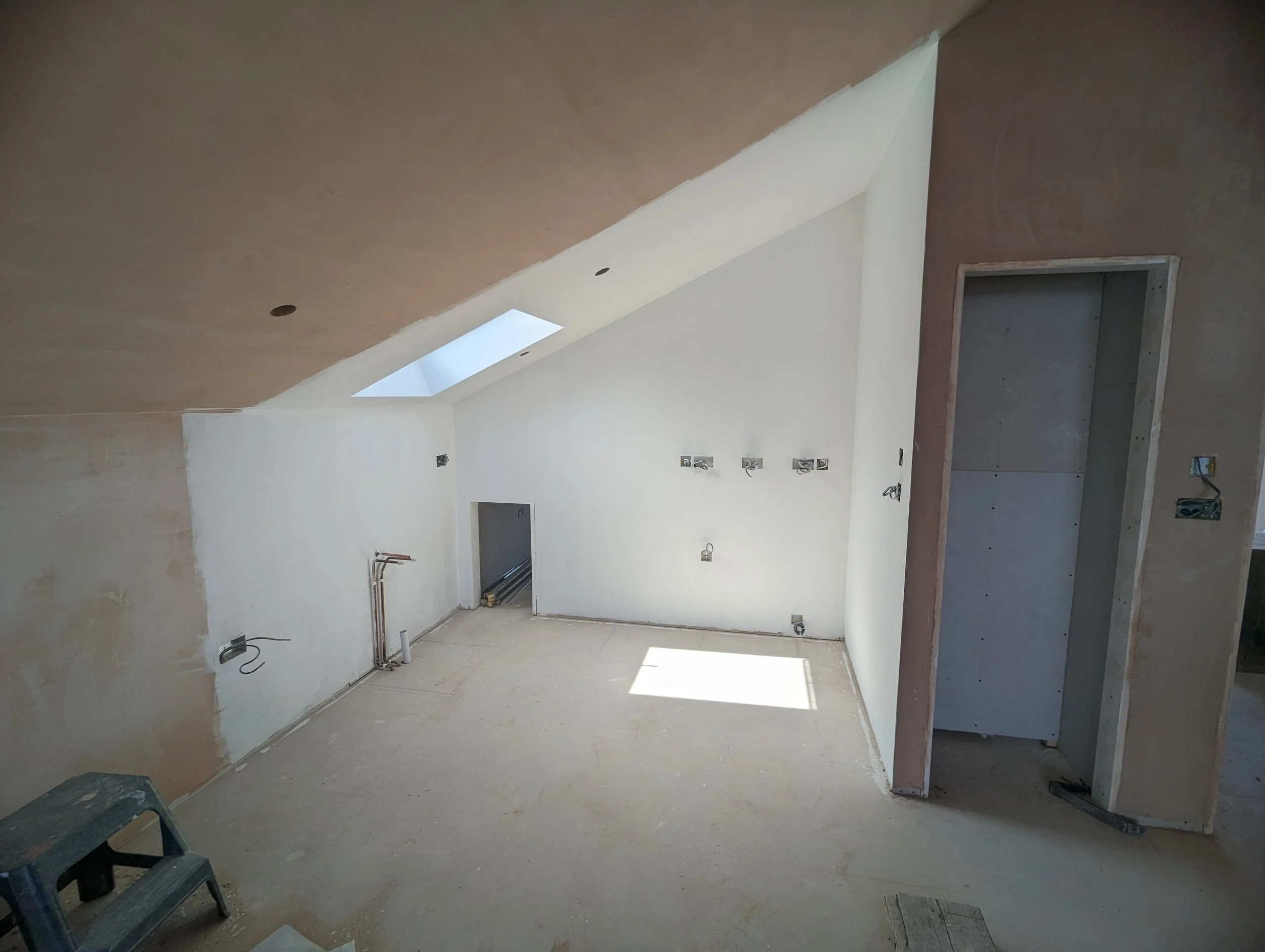Room under construction with white walls, a skylight, and exposed wiring and plumbing.