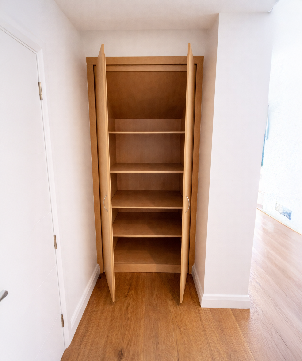 Empty wooden wardrobe with double doors next to a white wall inside a room with hardwood flooring.