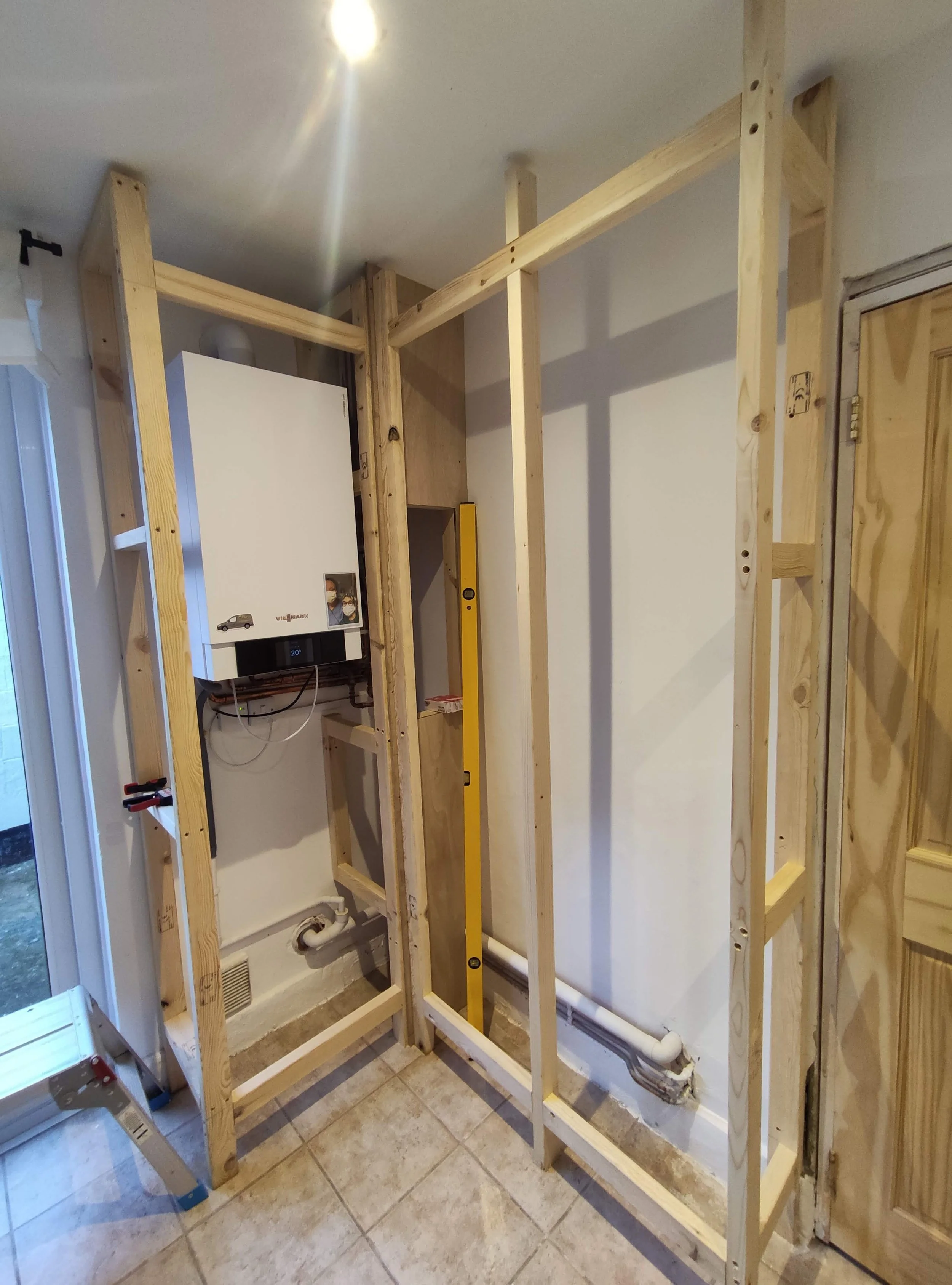 Construction frame for a wall in a room, with a boiler and plumbing pipes installed, and a level leaning against the framing.