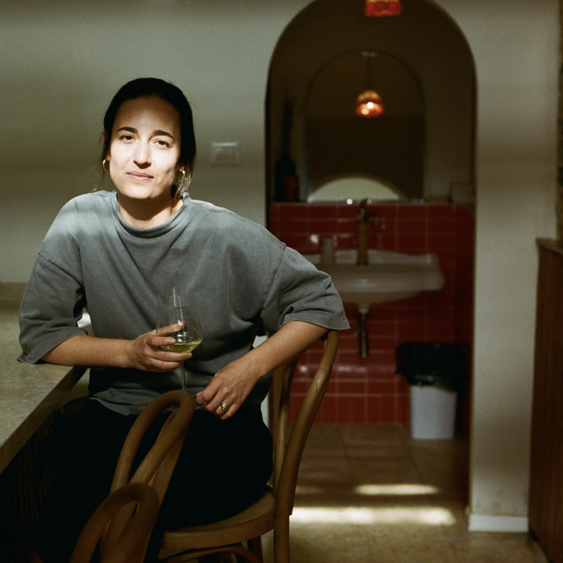 A woman sitting at a kitchen counter holding a wine glass with white wine, wearing a gray oversized t-shirt, in a cozy kitchen with warm lighting.