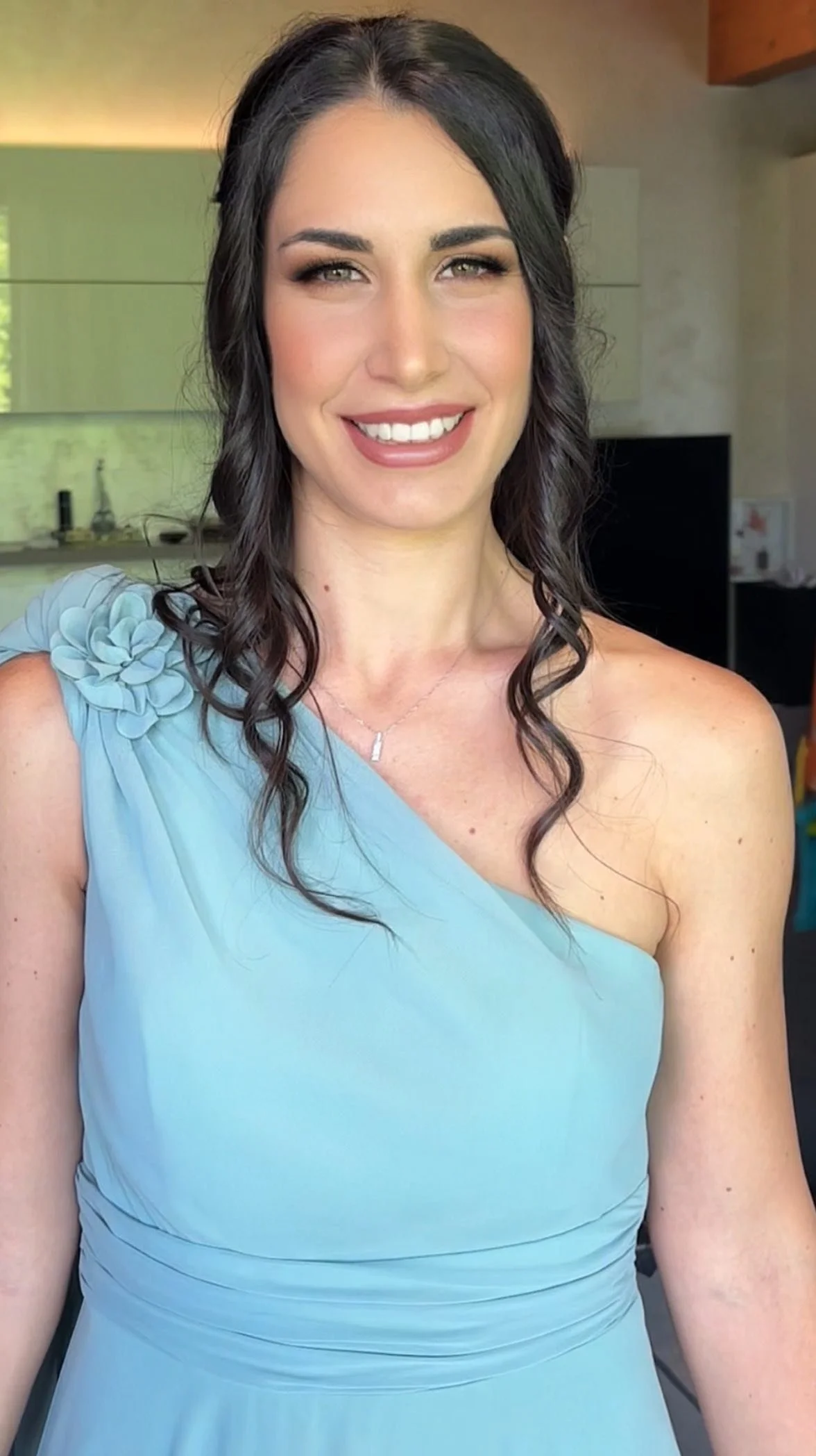 Donna sorridente con capelli ricci castani e abito azzurro con dettaglio a fiore sulla spalla, in un ambiente casalingo.