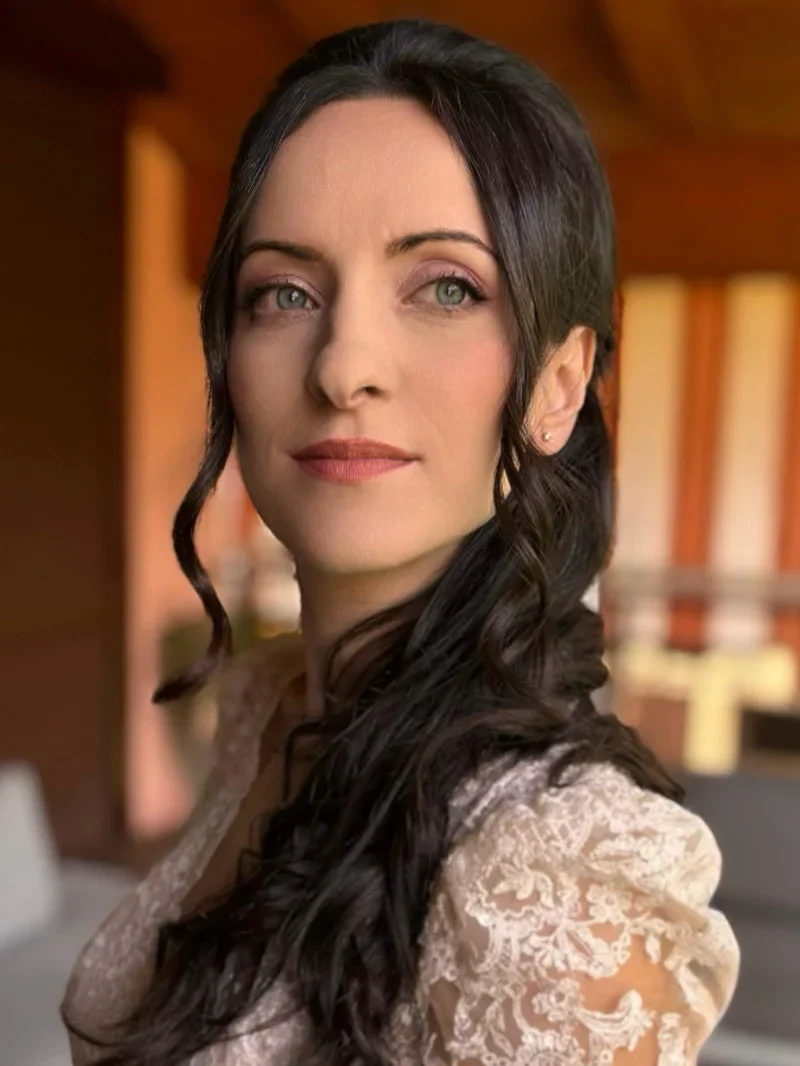 Ritratto di una donna con capelli lunghi ricci scuri, occhi azzurri e la bocca leggermente sorridente, indossa un vestito in pizzo cream.