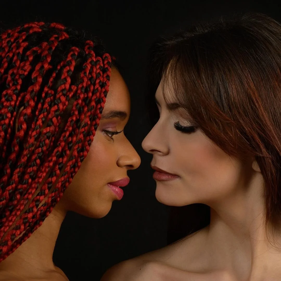 Due donne si fronteggiano con i volti molto vicini, occhi chiusi, sfondo nero.
