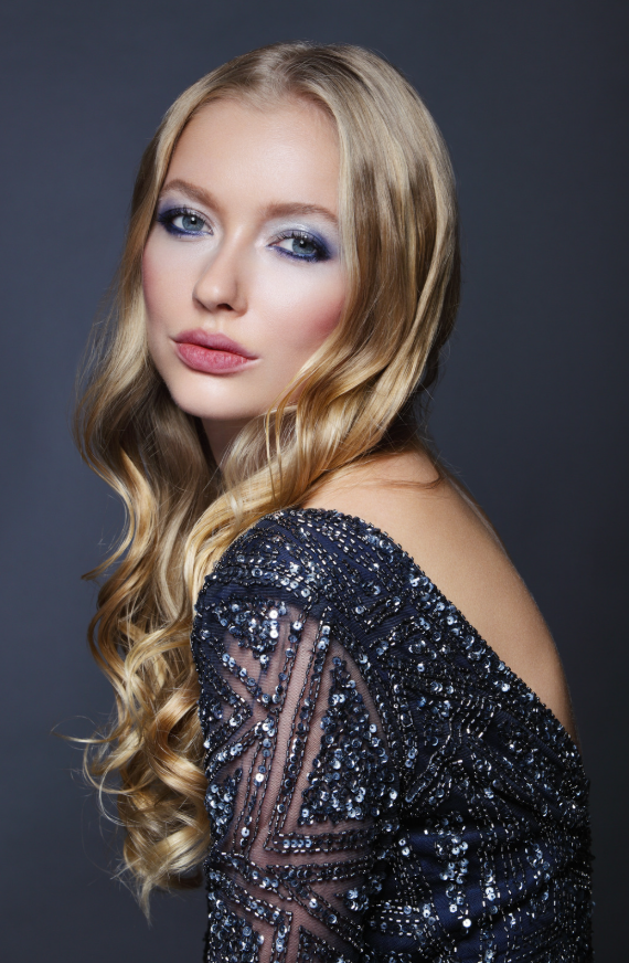 Ragazza con trucco occhi blu, abbigliamento nero decorato con paillettes, capelli lunghi ondulati, sfondo grigio scuro.