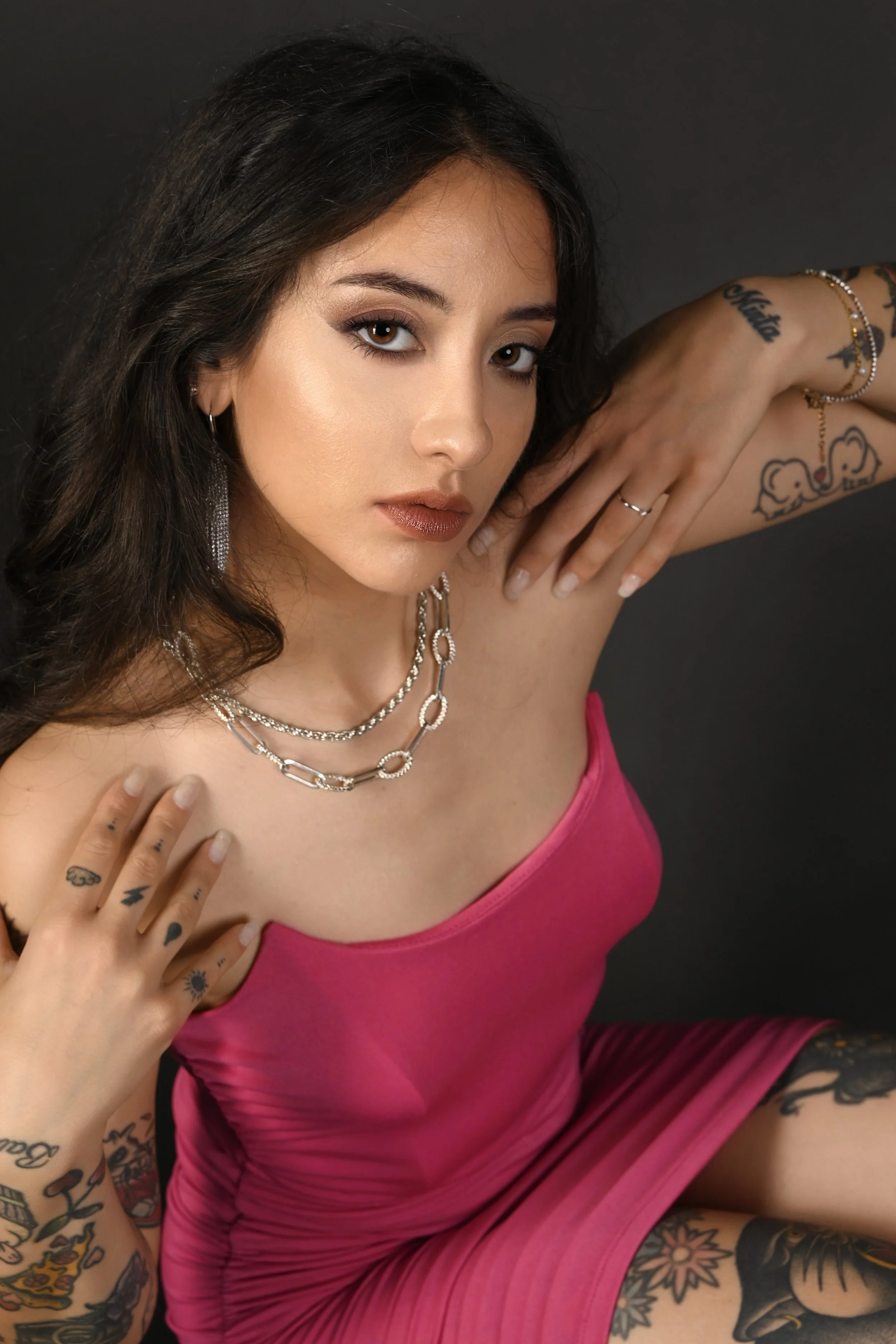 Ragazza con tatuaggi, in abito rosa, che indossa orecchini e collane, con trucco naturale, posa con braccia sollevate e sfondo nero.