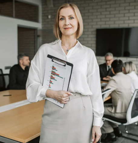 Una donna sorridente in abbigliamento formale che tiene una cartella con grafici e dati, in una sala conferenze con altre persone al tavolo.