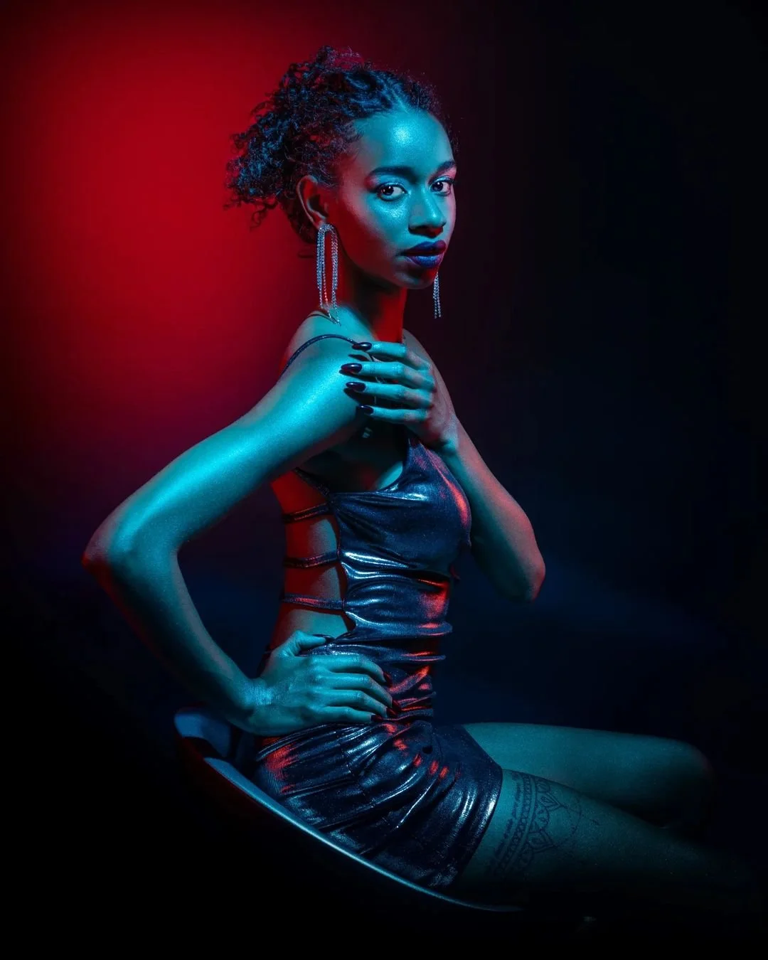 Ragazza con acconciatura riccia, abbigliamento lucido, orecchini pendenti, posa sensuale, illuminata da luci blu e rosse