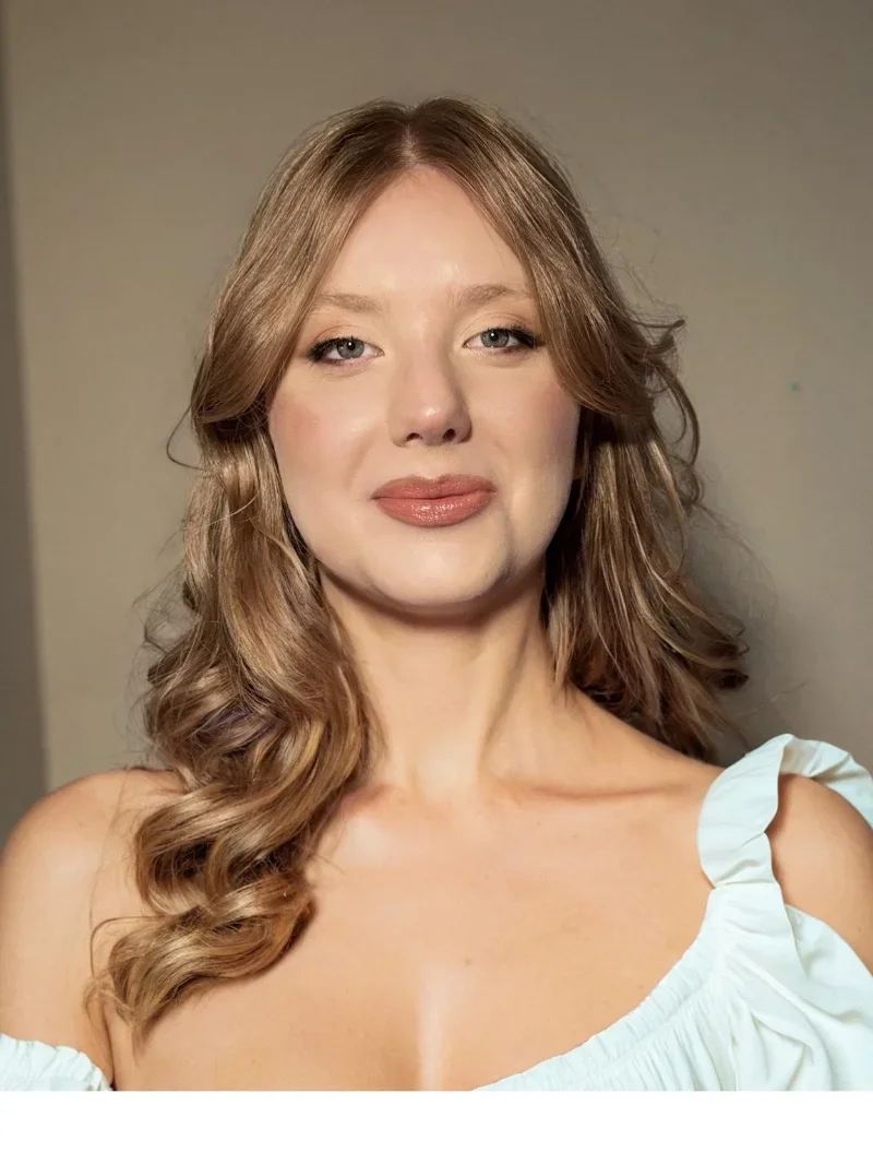  Ritratto di una donna con capelli castani riccioli, trucco naturale e indossa una maglietta bianca con spalla scoperta.