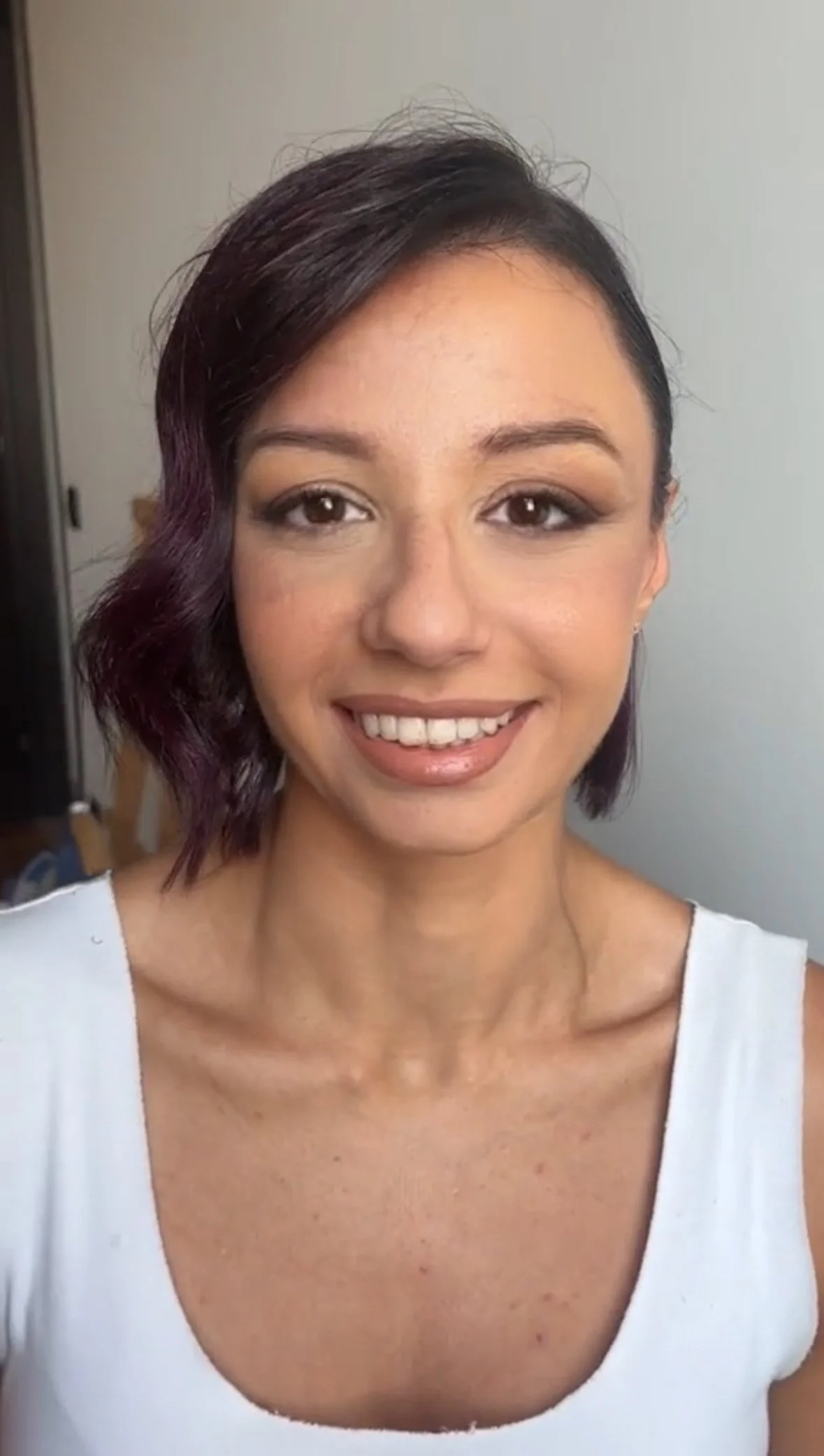  Ritratto di una donna con capelli corti neri e viola, sorriso, vestito bianco, sfondo neutro.